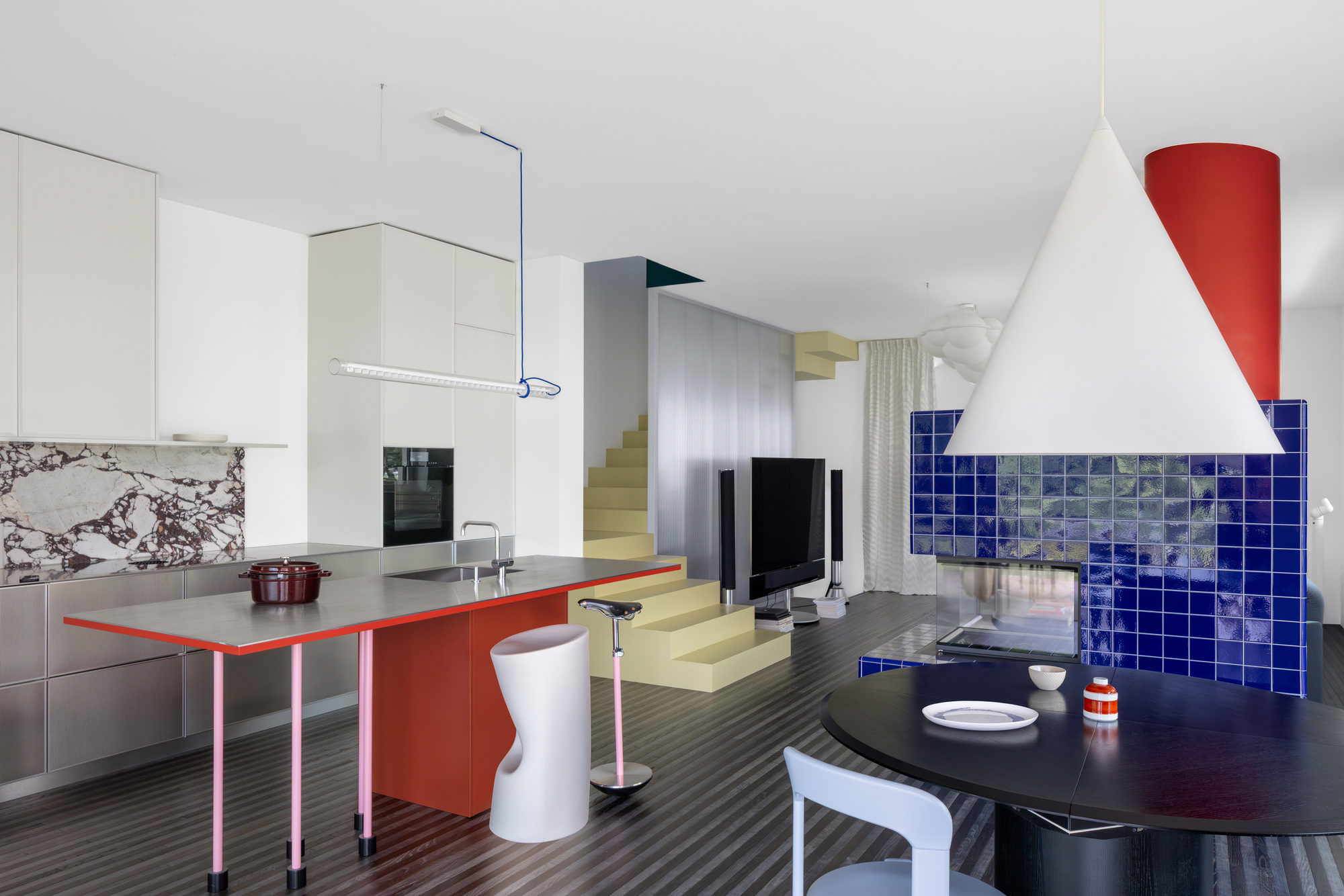 Open living space with a red-edged steel kitchen island, yellow stepped stair and cobalt tiled fireplace under a white cone pendant