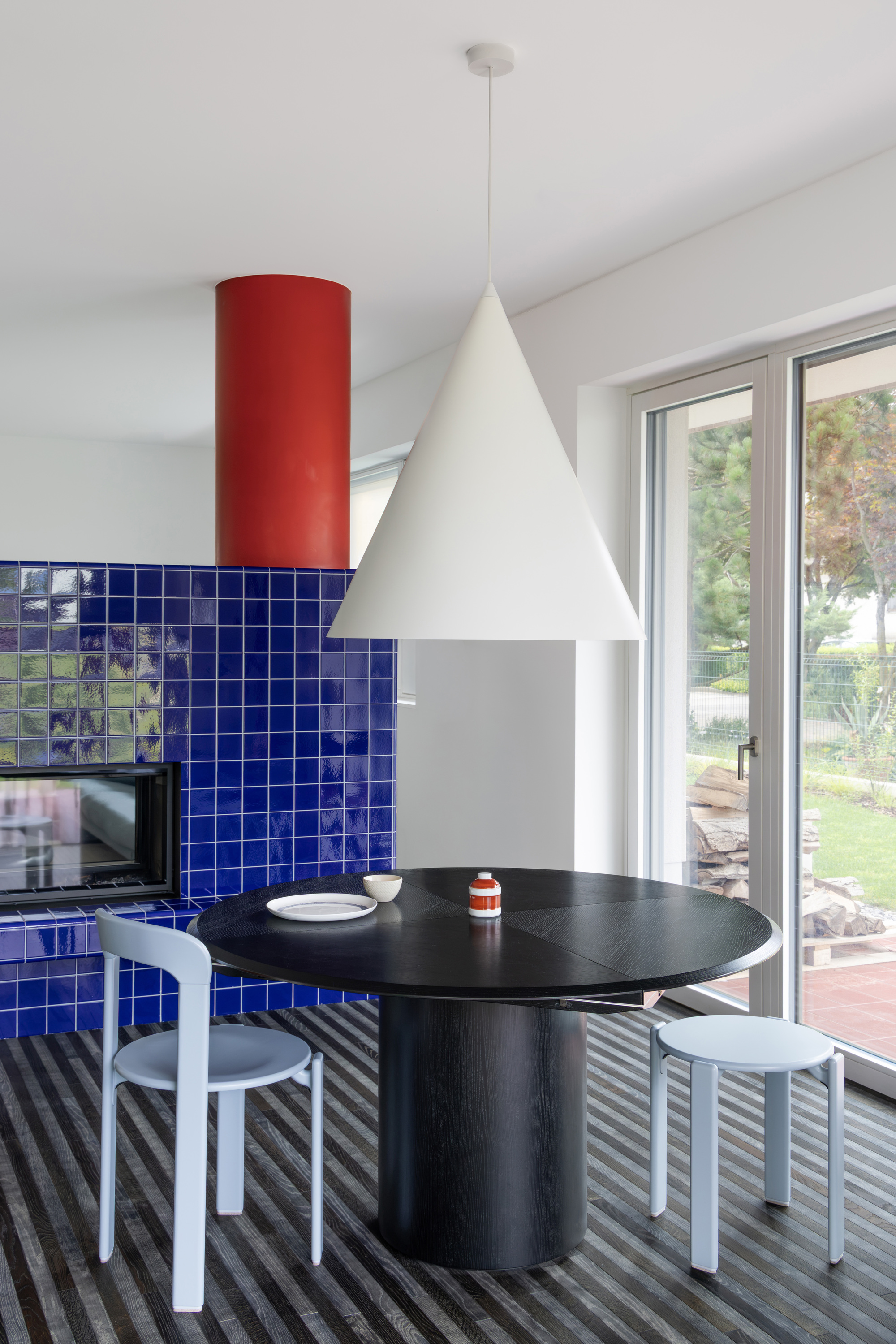 Round black dining table beneath a white cone pendant, with a blue tiled fireplace and garden glimpsed through glazed doors