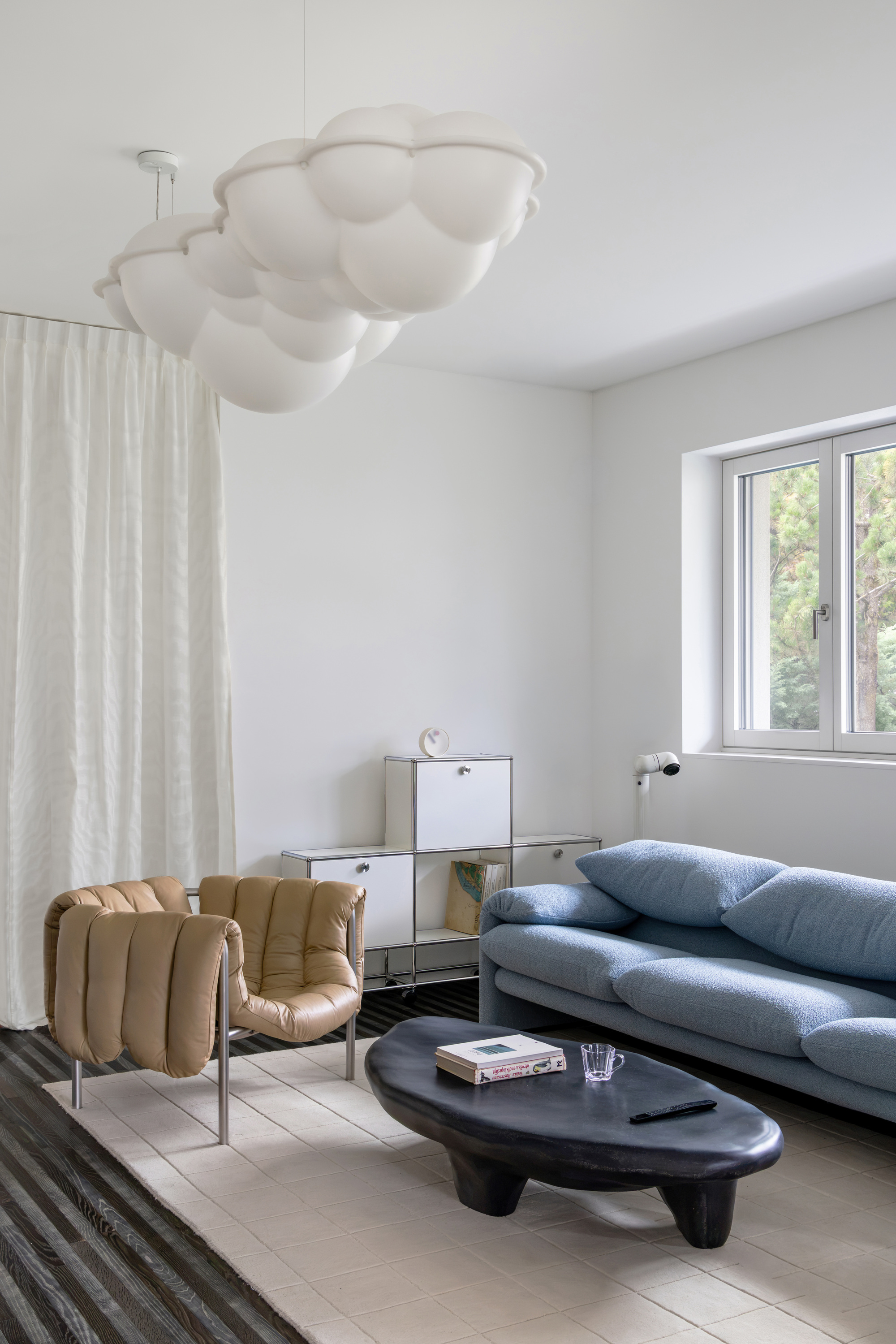 Seating corner with a blue sofa, tan quilted armchair and cloud-shaped pendant beside a sheer curtain and a window onto trees