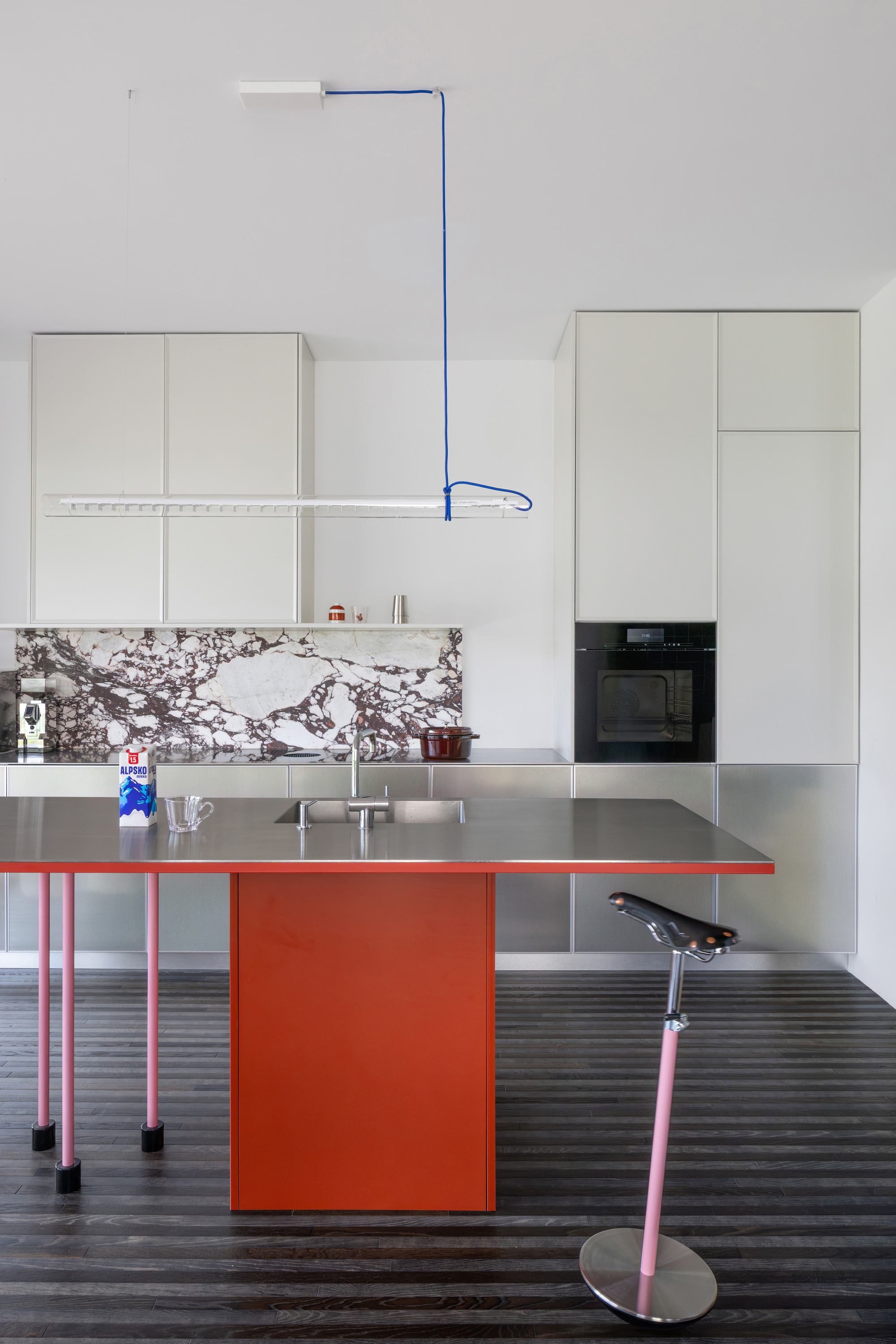 Kitchen with a red island, pink legs and a marble splashback, lit by a linear pendant hung from a blue cord