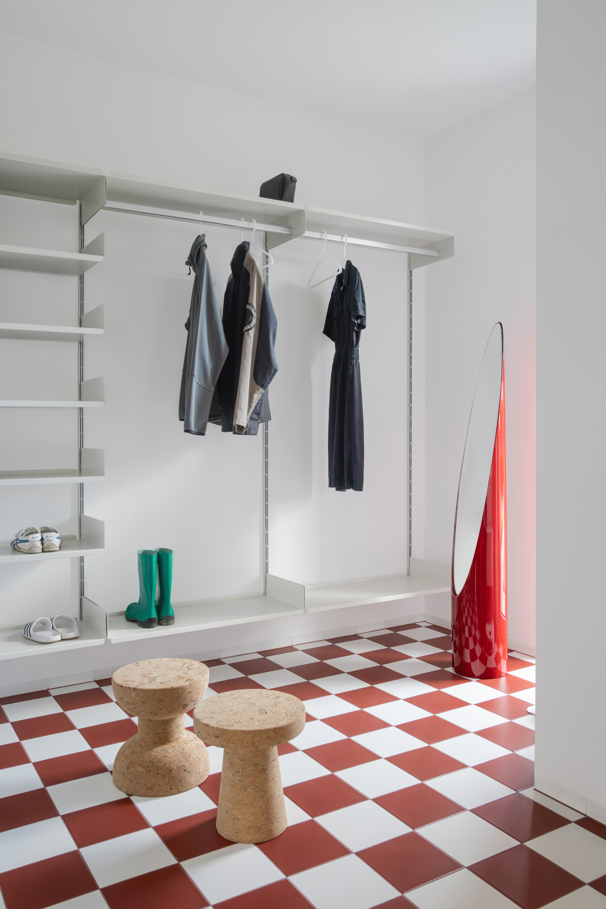 Entry alcove with open wall shelving and hanging jackets, cork stools and a red mirror on a red and white checkered floor