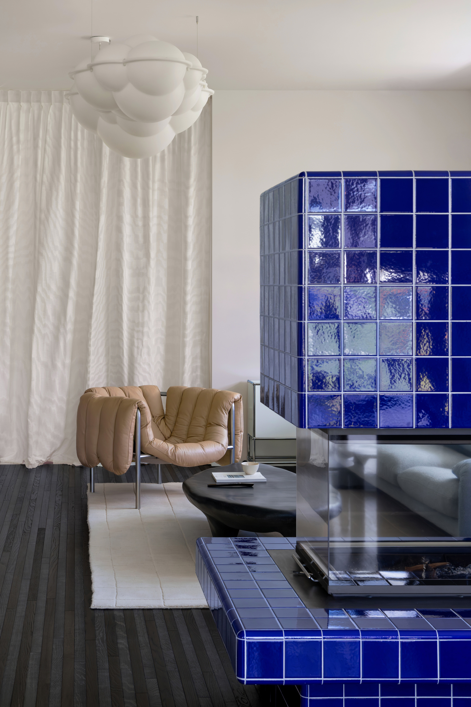 Cobalt glazed tile fireplace volume in the foreground, with a tan armchair, black coffee table and sheer curtain behind in soft daylight