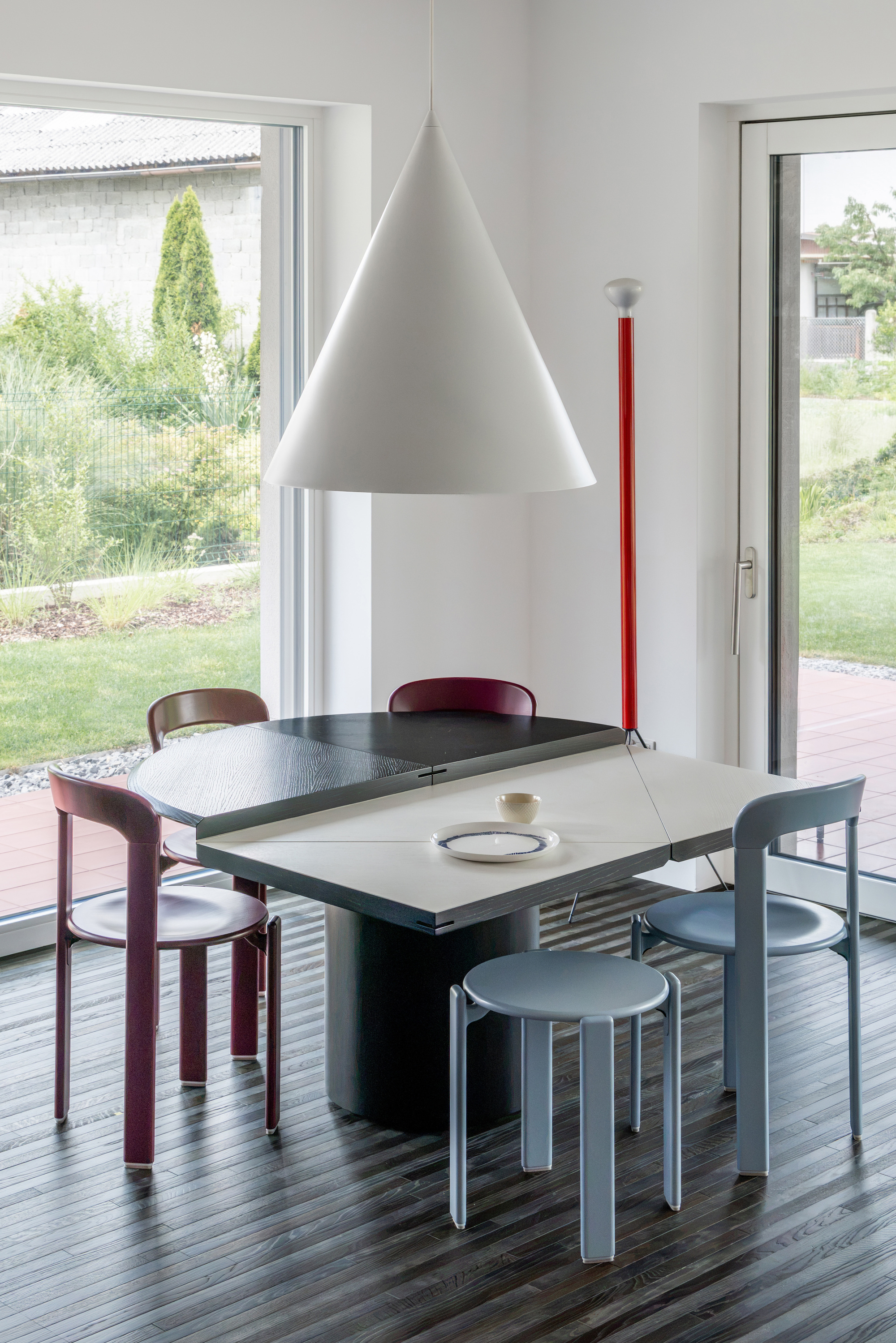 Dining corner with a white cone pendant over a split-top table, maroon and blue chairs, and garden views through glass