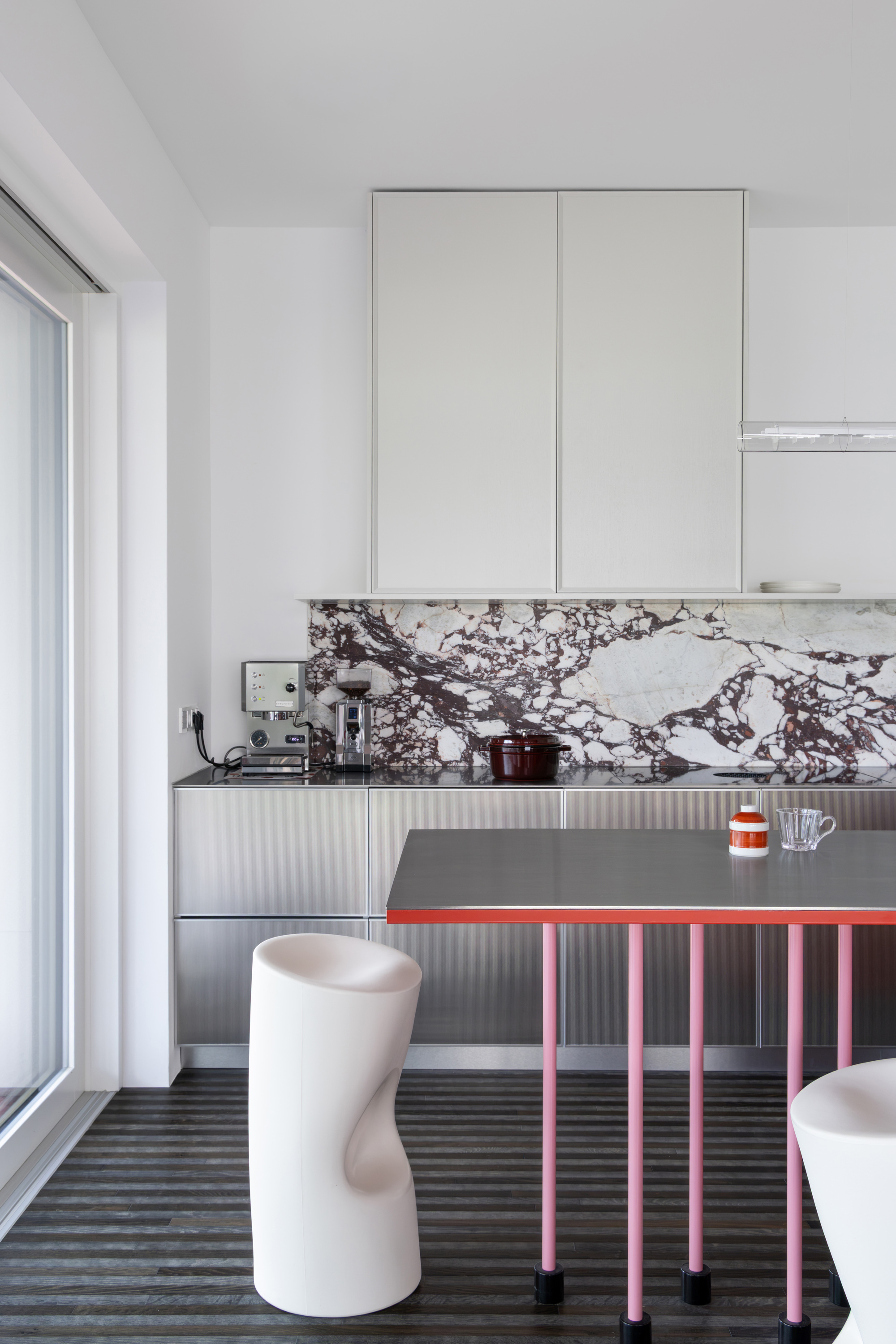 Kitchen with stainless steel cabinets, marble splashback and a pink-legged table, lit by daylight from a sliding glass door