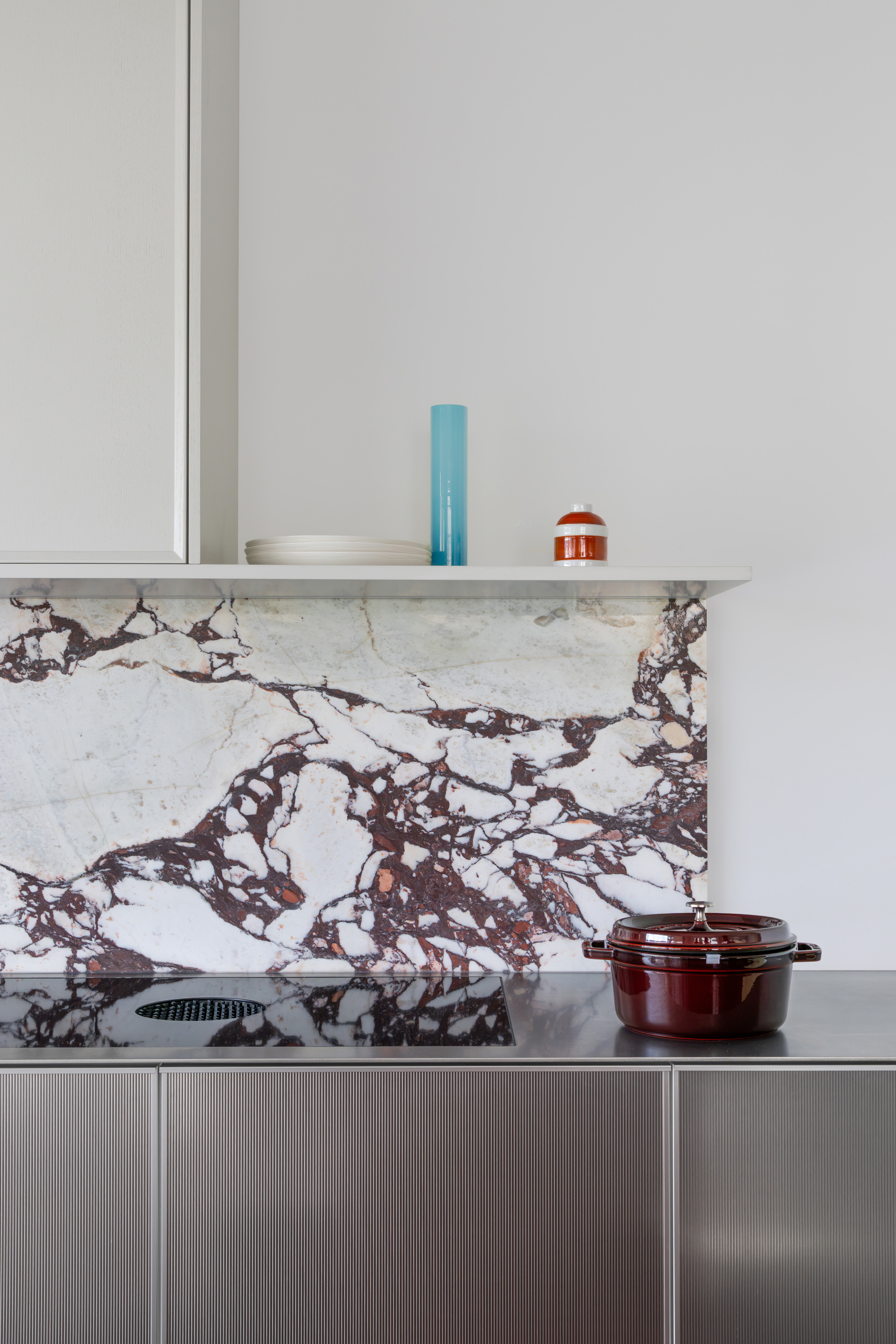 Veined marble splashback beneath a floating shelf holding plates, a blue glass vase and a red enamel pot on the counter