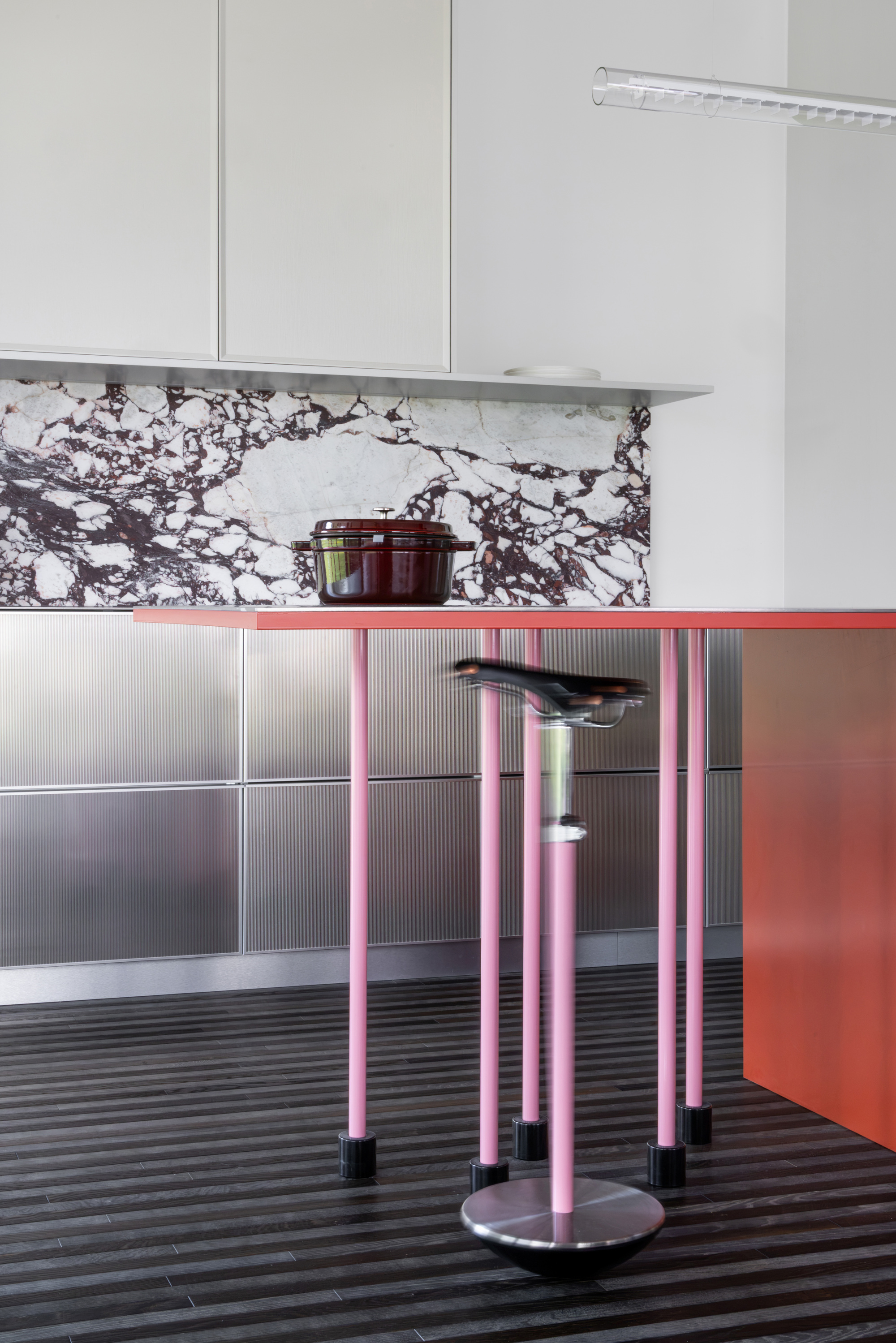 Pink tubular table legs with black feet beside a tilting stool, stainless cabinets and marble backsplash behind in soft daylight