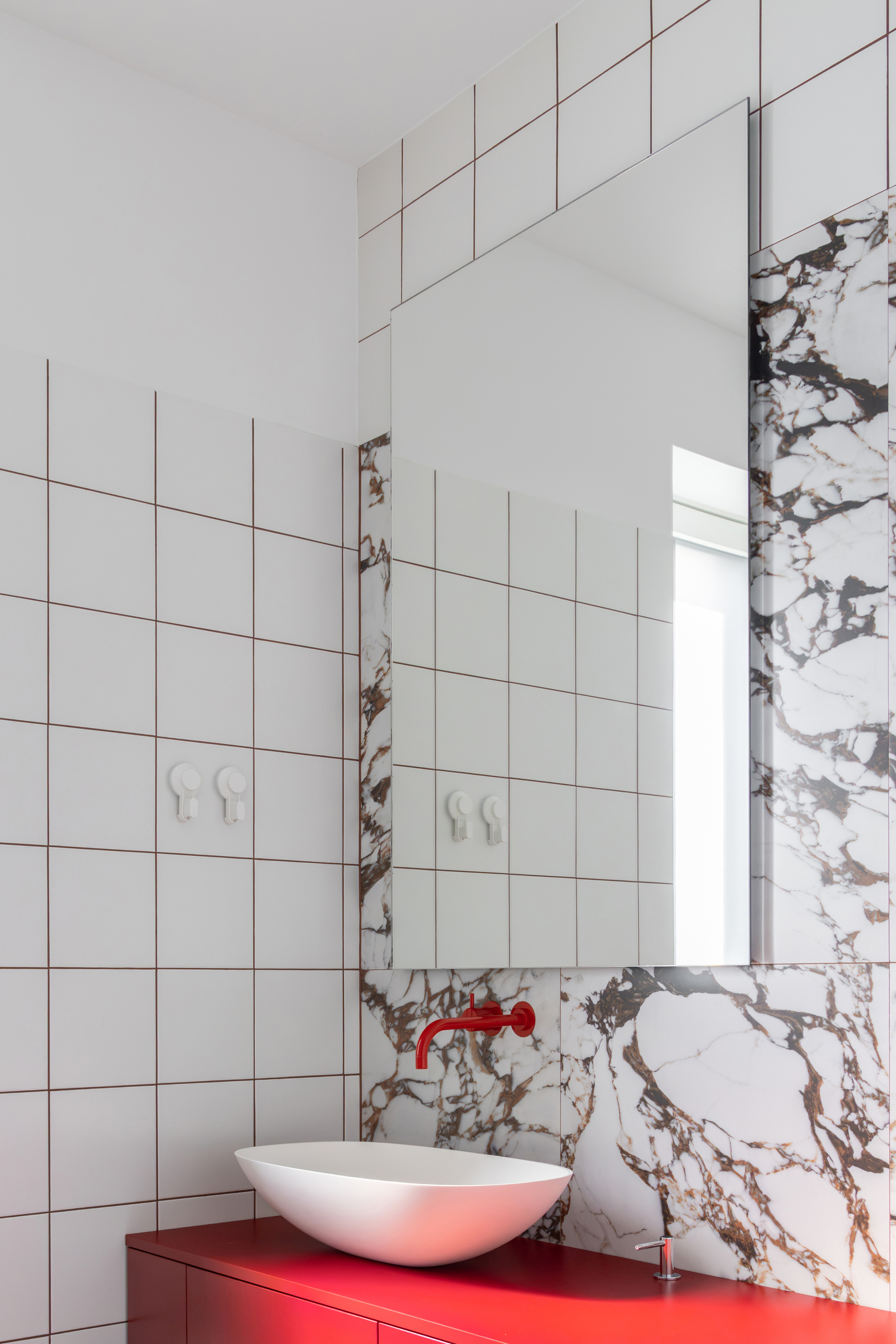 Bathroom corner with a white vessel basin, red wall-mounted tap and red vanity against square white tiles and veined marble