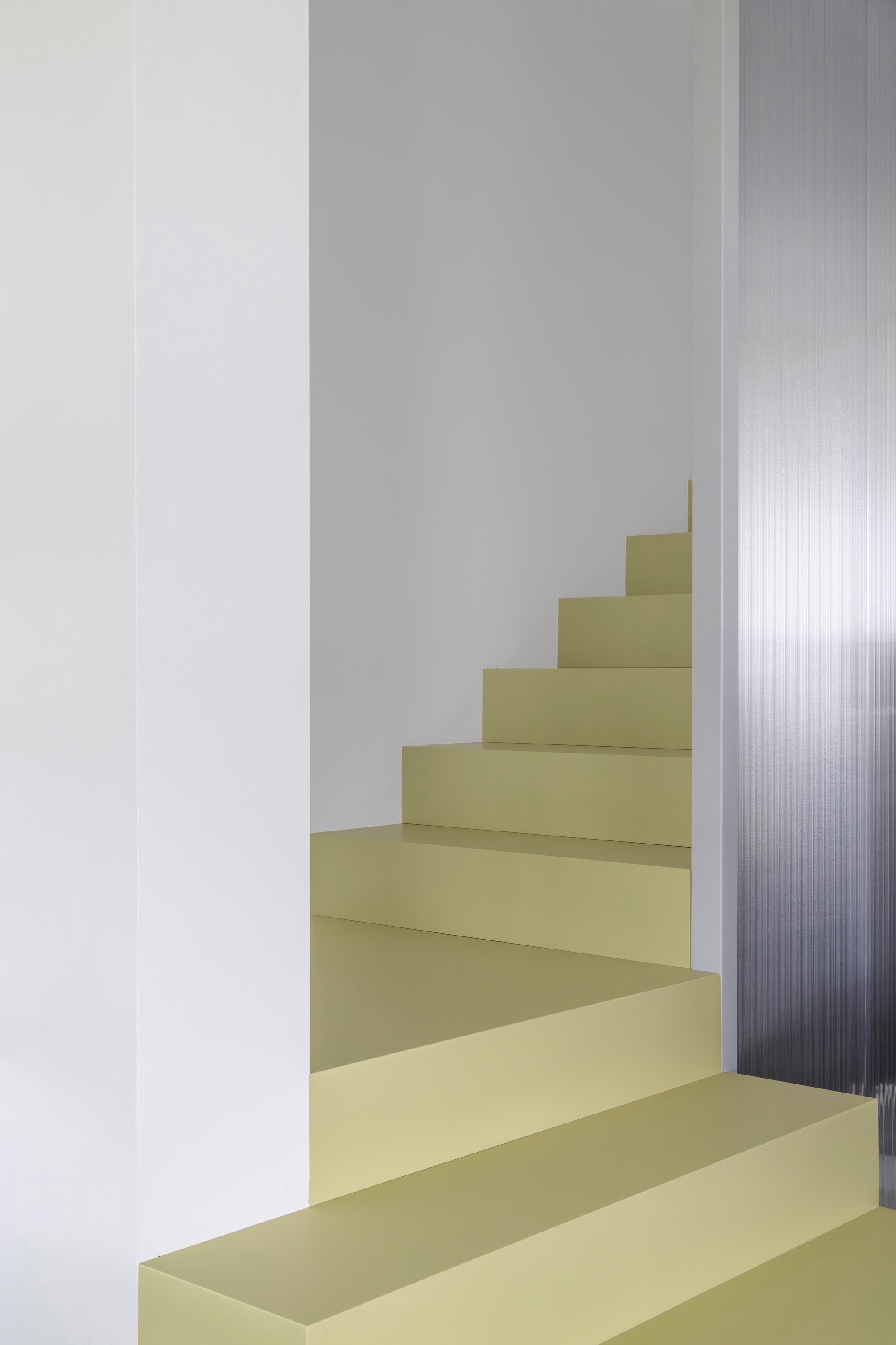 Pale olive stair turning through a white stairwell beside a ribbed translucent polycarbonate panel in soft daylight