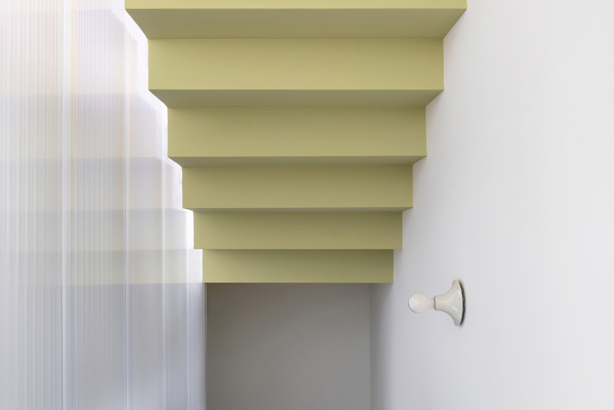 Underside of the olive stair treads seen from below, beside a ribbed translucent screen and a white ceramic wall lamp