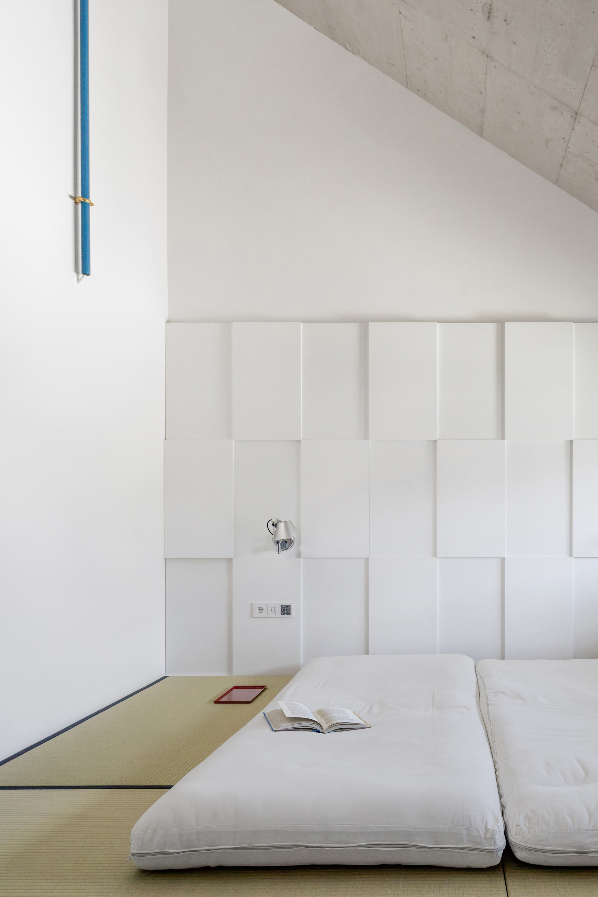 Bedroom with white floor mattresses on tatami mats below a staggered white panel headboard and raw concrete sloping ceiling