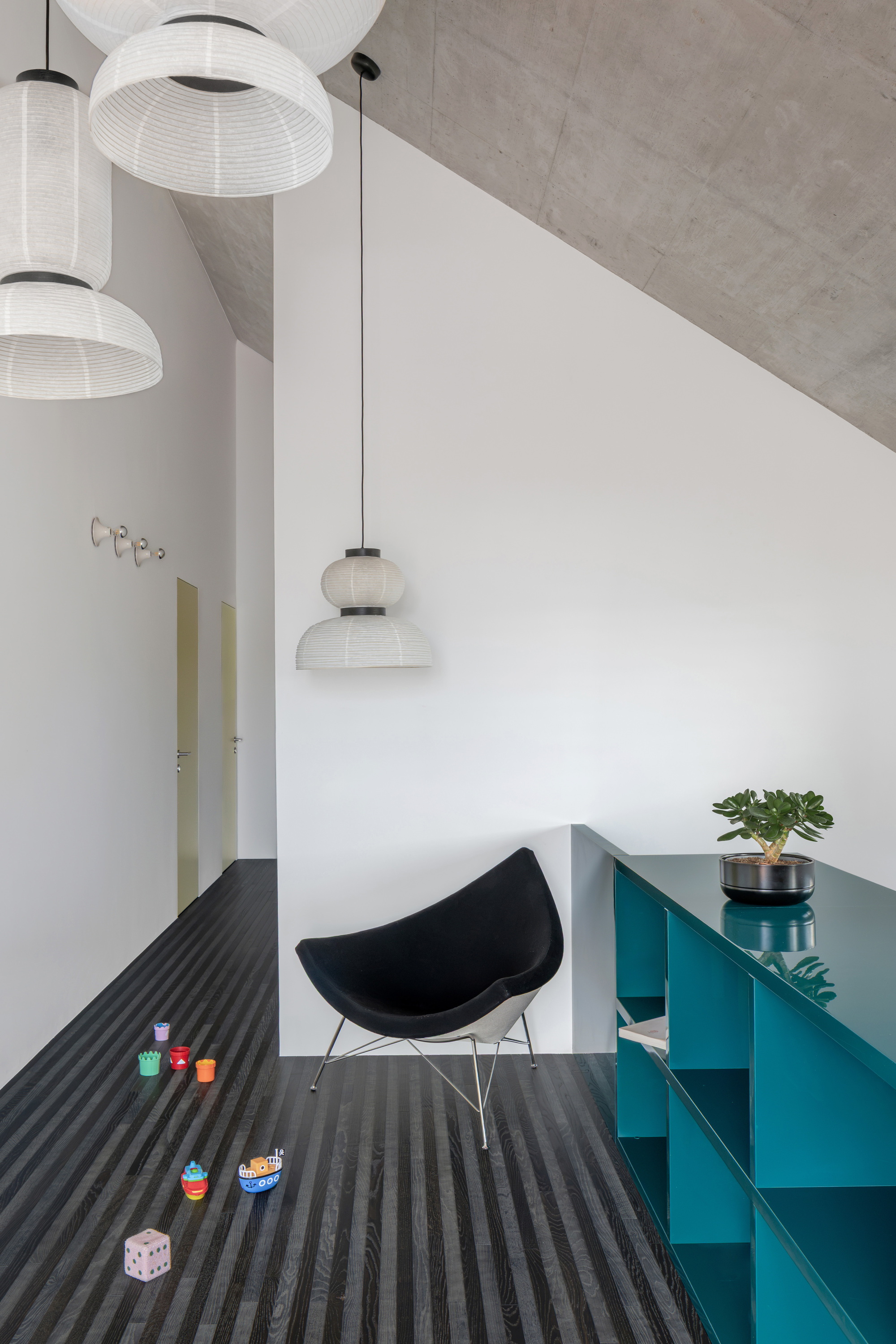 Black lounge chair beside a teal shelving unit under paper pendant lamps, with toys scattered on the striped dark floor