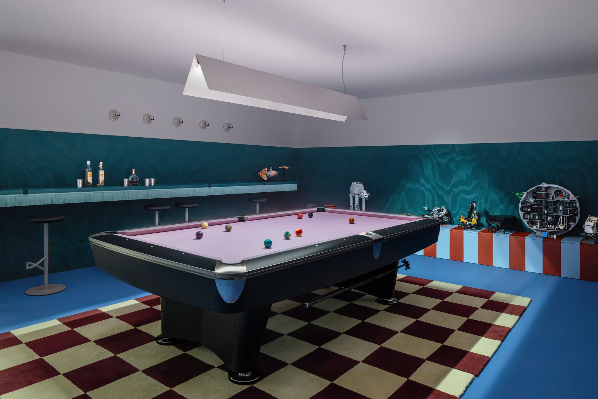 Games room with a lilac-felt pool table on a red and cream checkered rug, teal wood-panelled walls and a bar shelf
