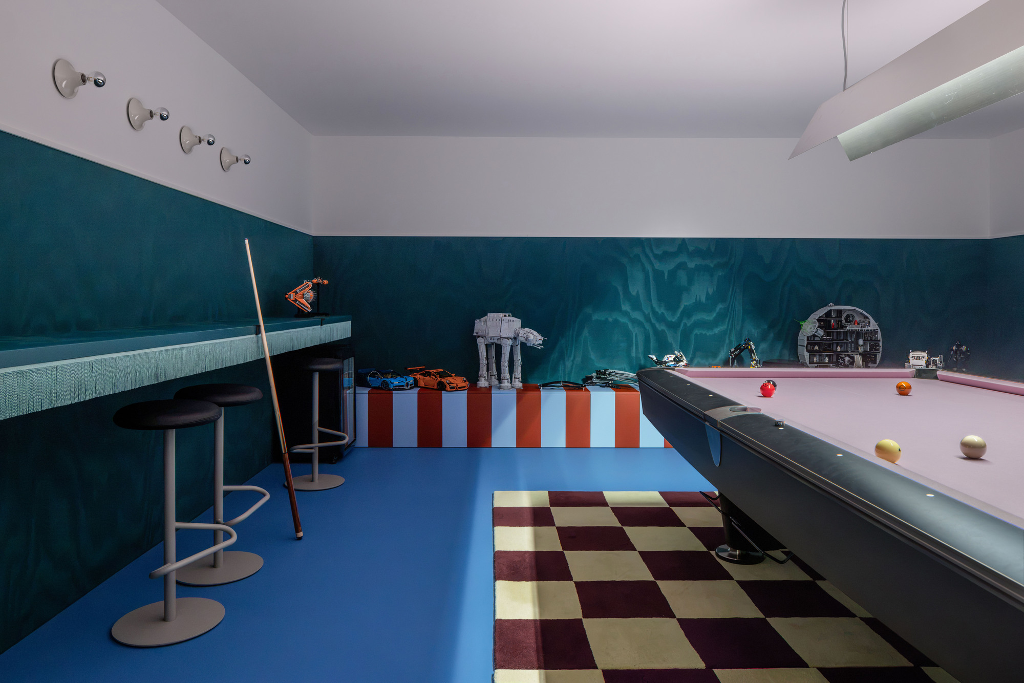 Games room with a pool table, teal plywood walls, striped display bench and checkered rug on a bright blue floor