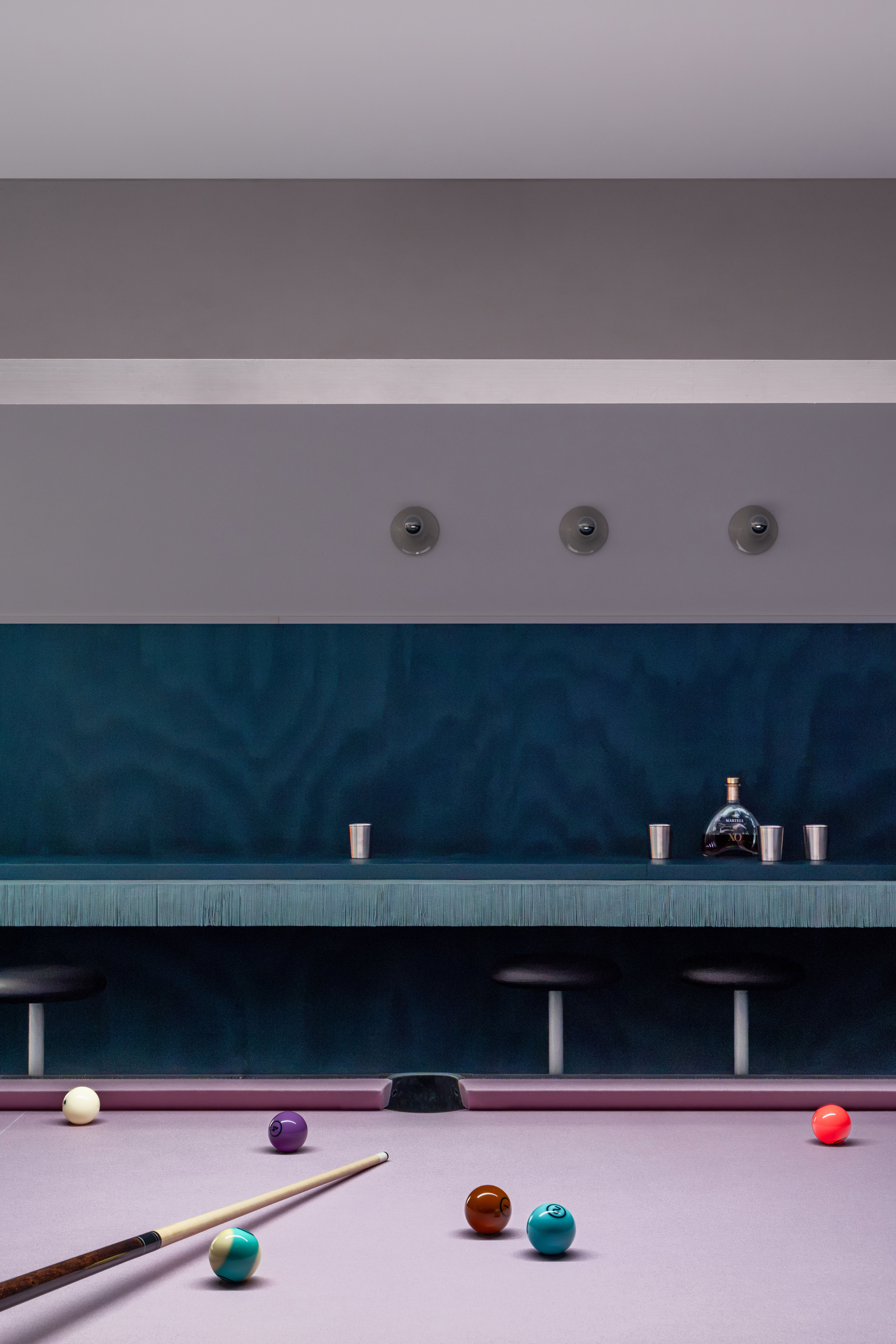 Pool table balls and a cue in the foreground, with a teal wall ledge holding a decanter and metal cups above bar stools