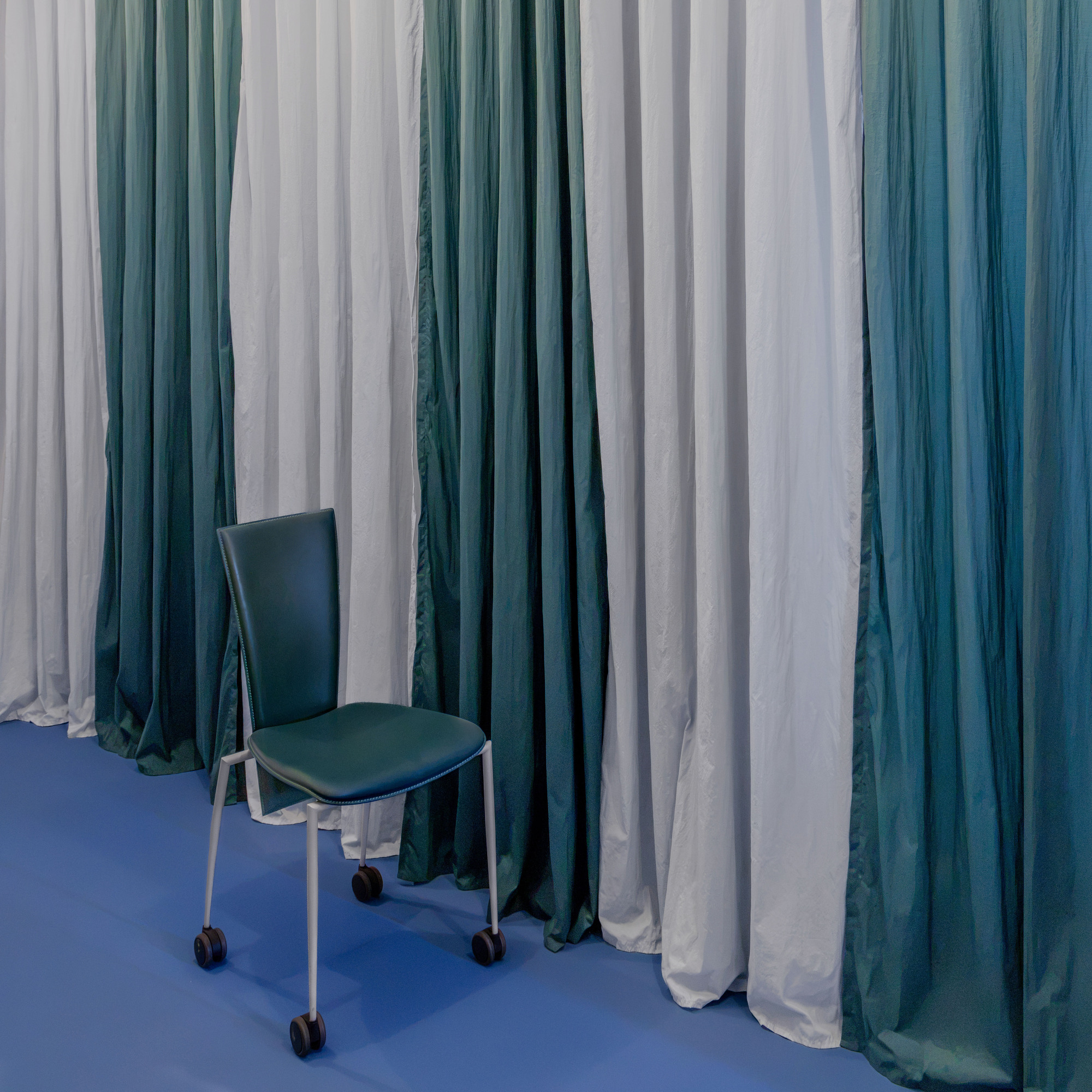 Green leather chair on casters in front of floor-length curtains alternating pale grey and teal, above a blue floor