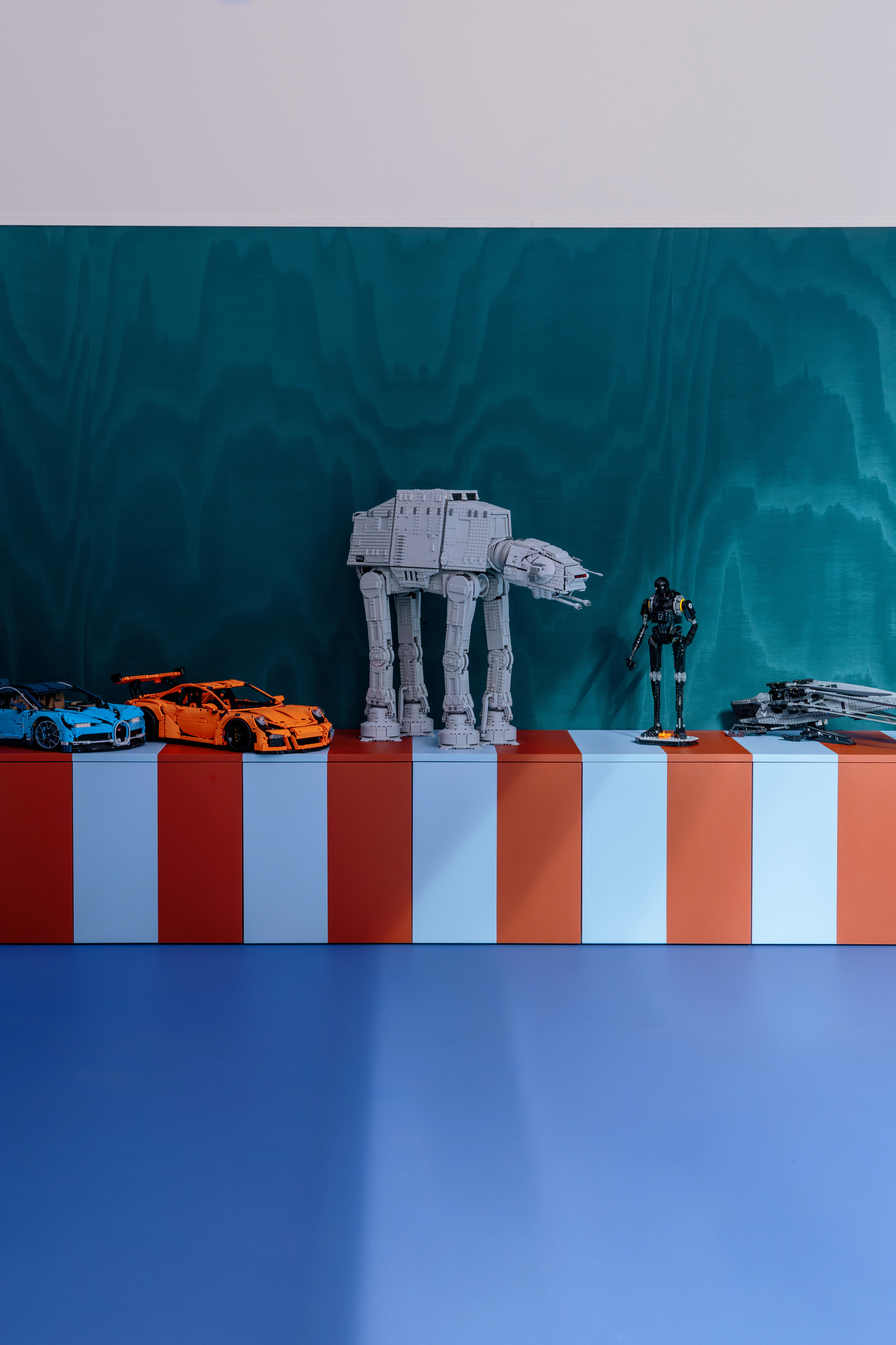 Red and pale blue striped bench displaying brick-built toy cars and a walker model against a teal wood-grain wall