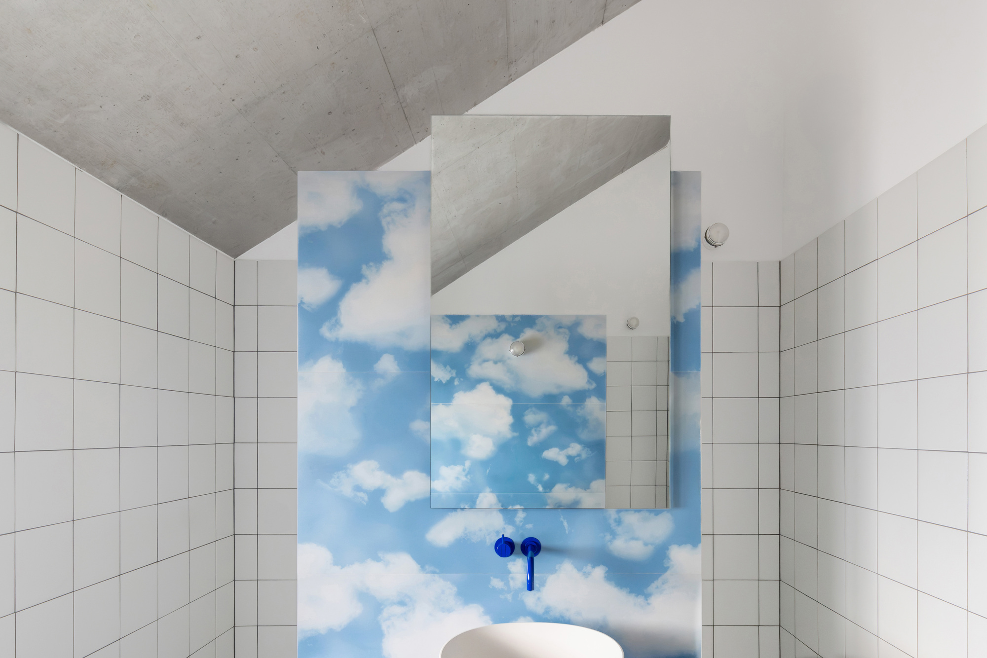 Bathroom basin wall printed with a cloud sky, blue wall tap and mirror, set under a sloping raw concrete ceiling