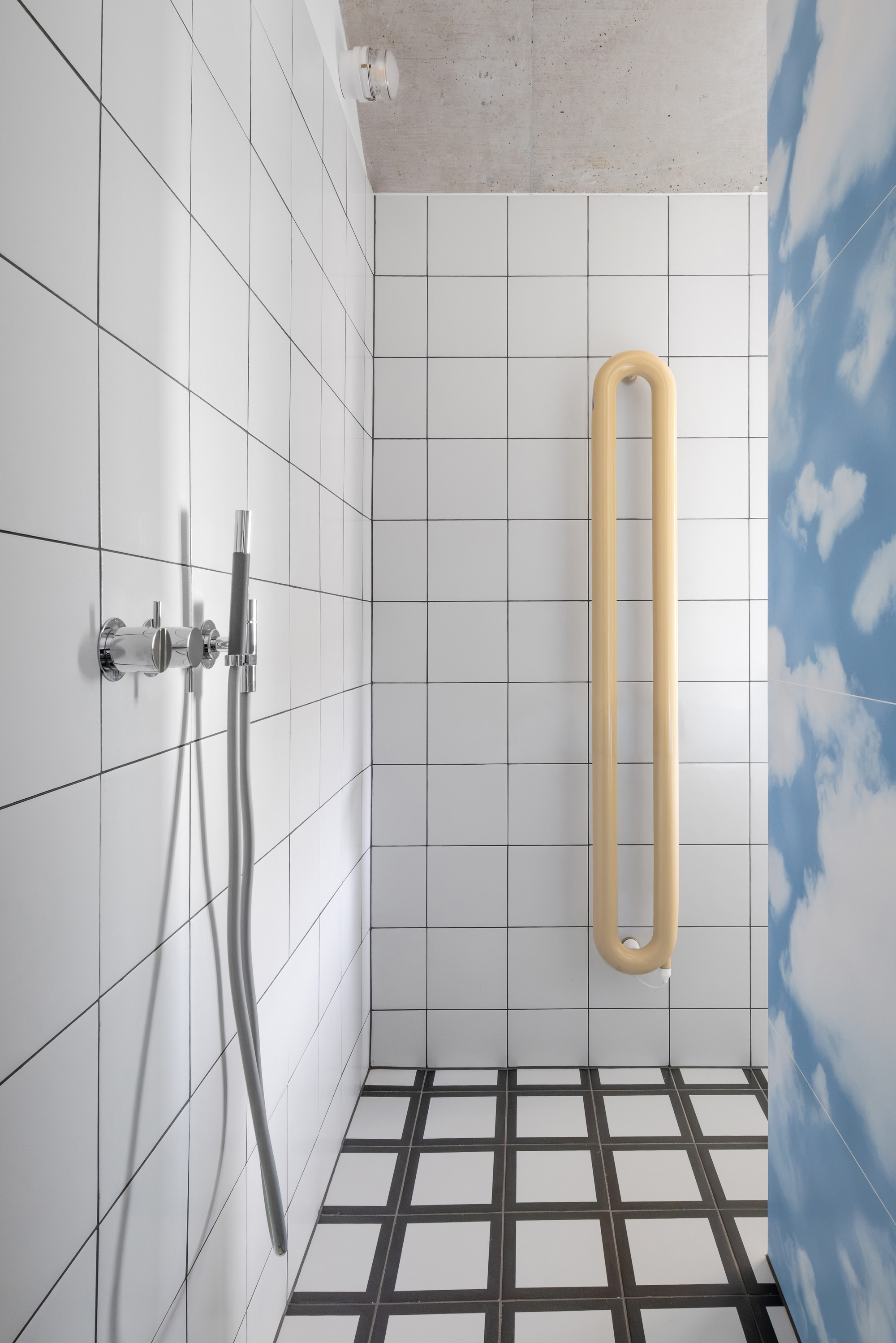 Shower corner with white square tiles, chrome handheld fixture, yellow looped towel radiator and black and white checked floor beside the cloud panel
