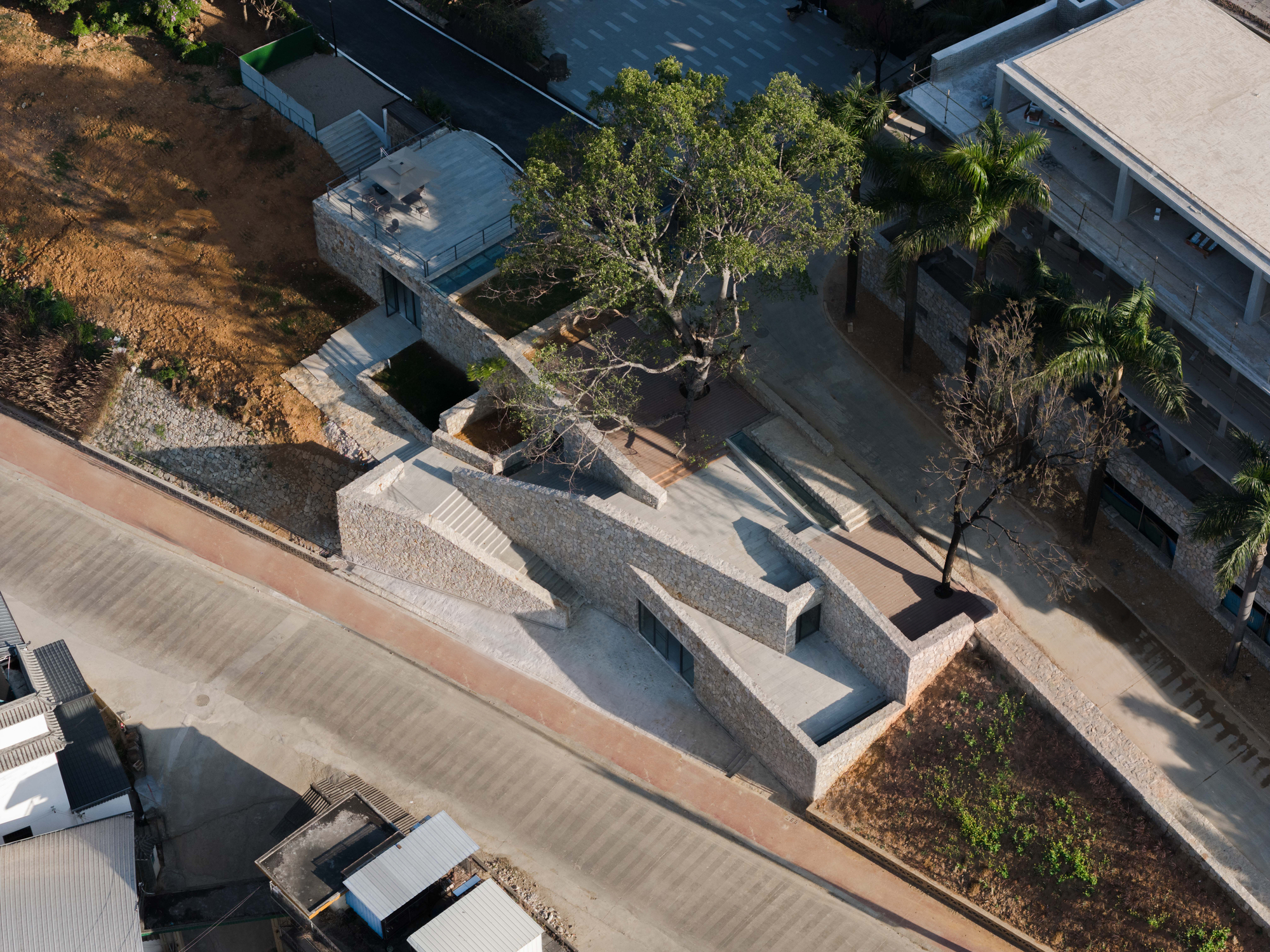 Aerial view of stepped rubble stone walls and rooftop terraces beside a road, with a large tree and palms nearby