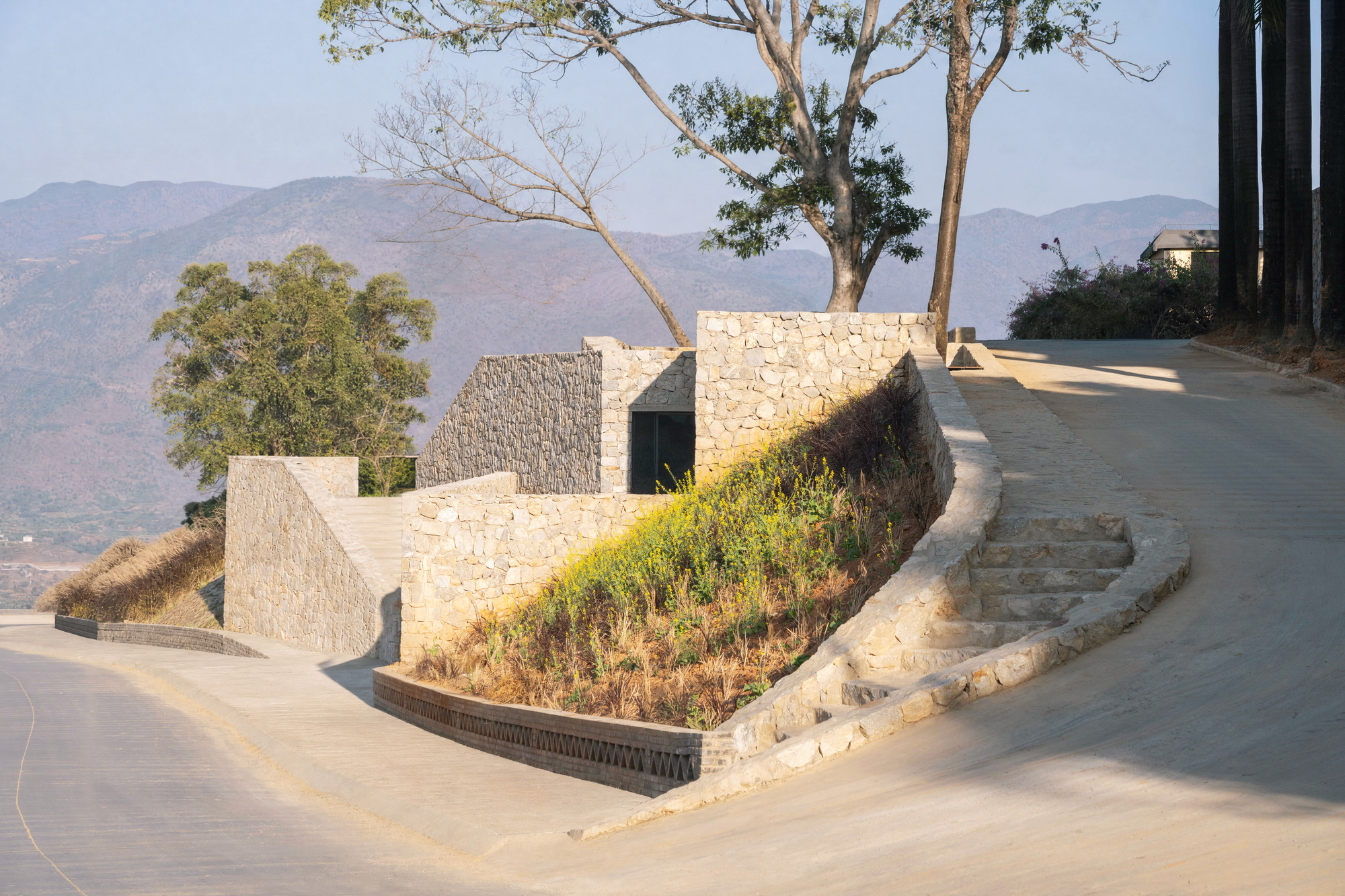 Curving stone retaining wall with a planted slope and stone steps beside a road, with hazy mountains behind in afternoon sun