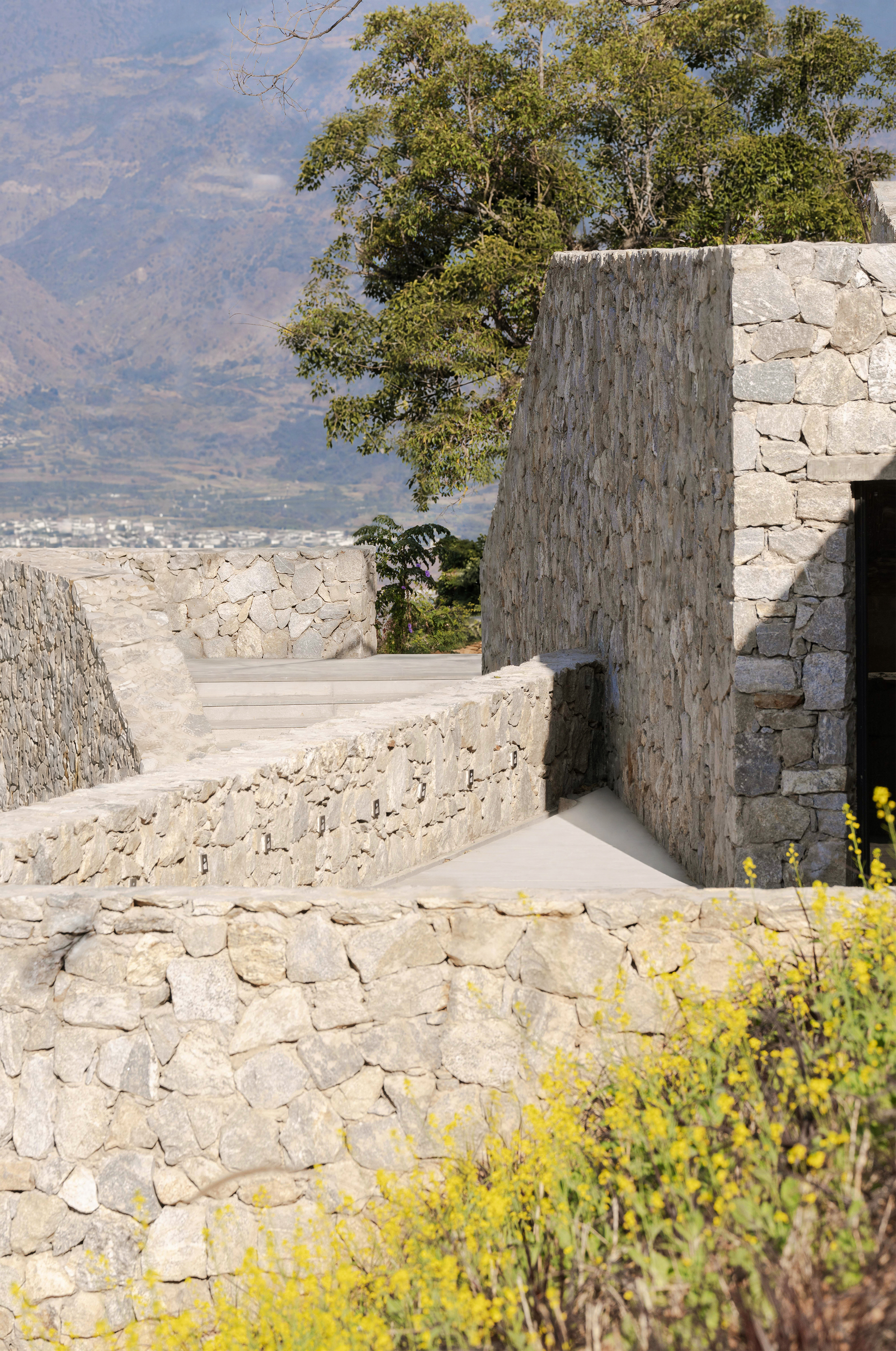 Close view of dry-laid stone walls and a concrete ramp, with yellow flowers in the foreground and a hillside beyond