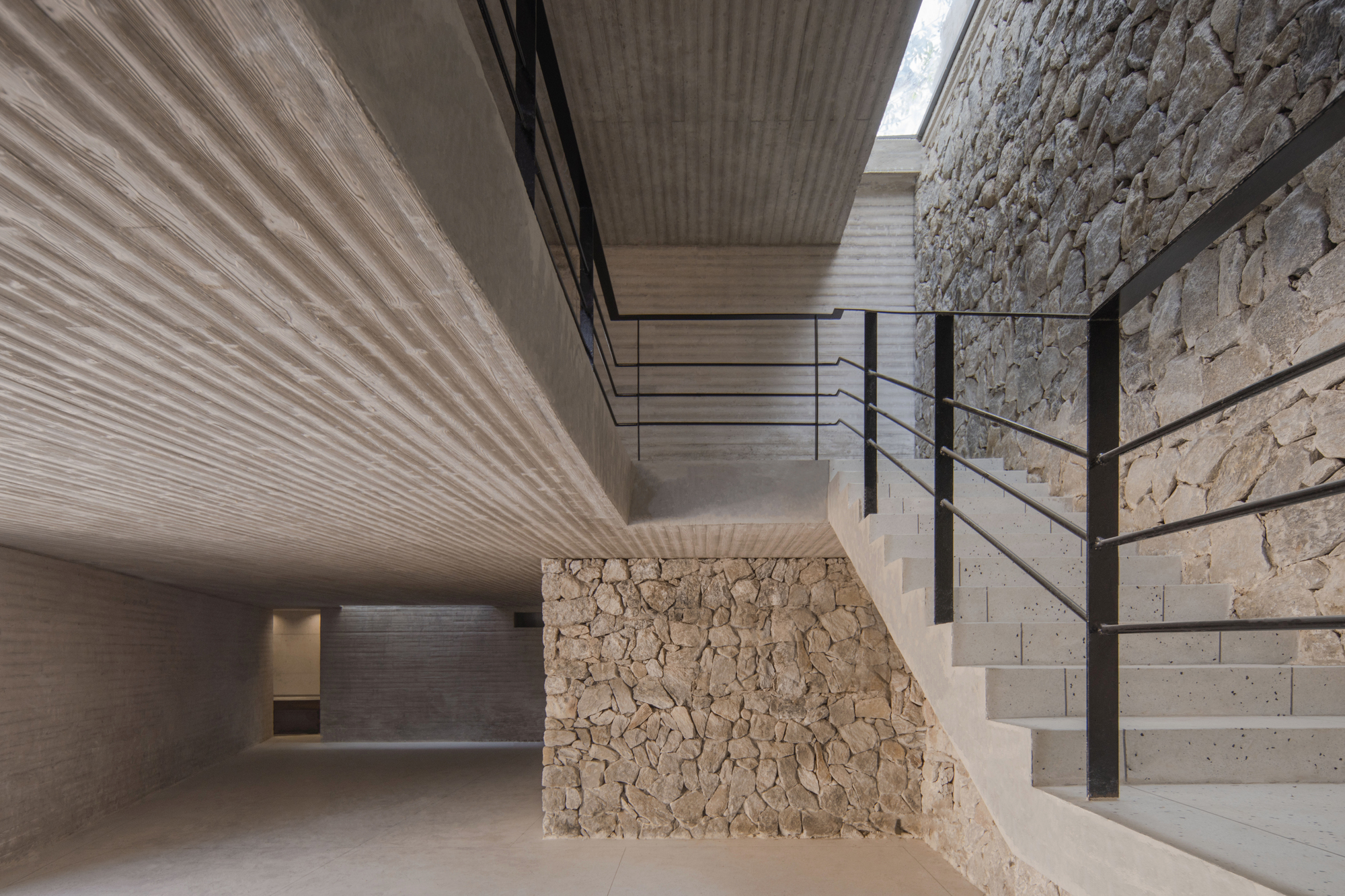 Interior stair with black steel railing between board-marked concrete soffits and rough stone walls, with daylight from a skylight above
