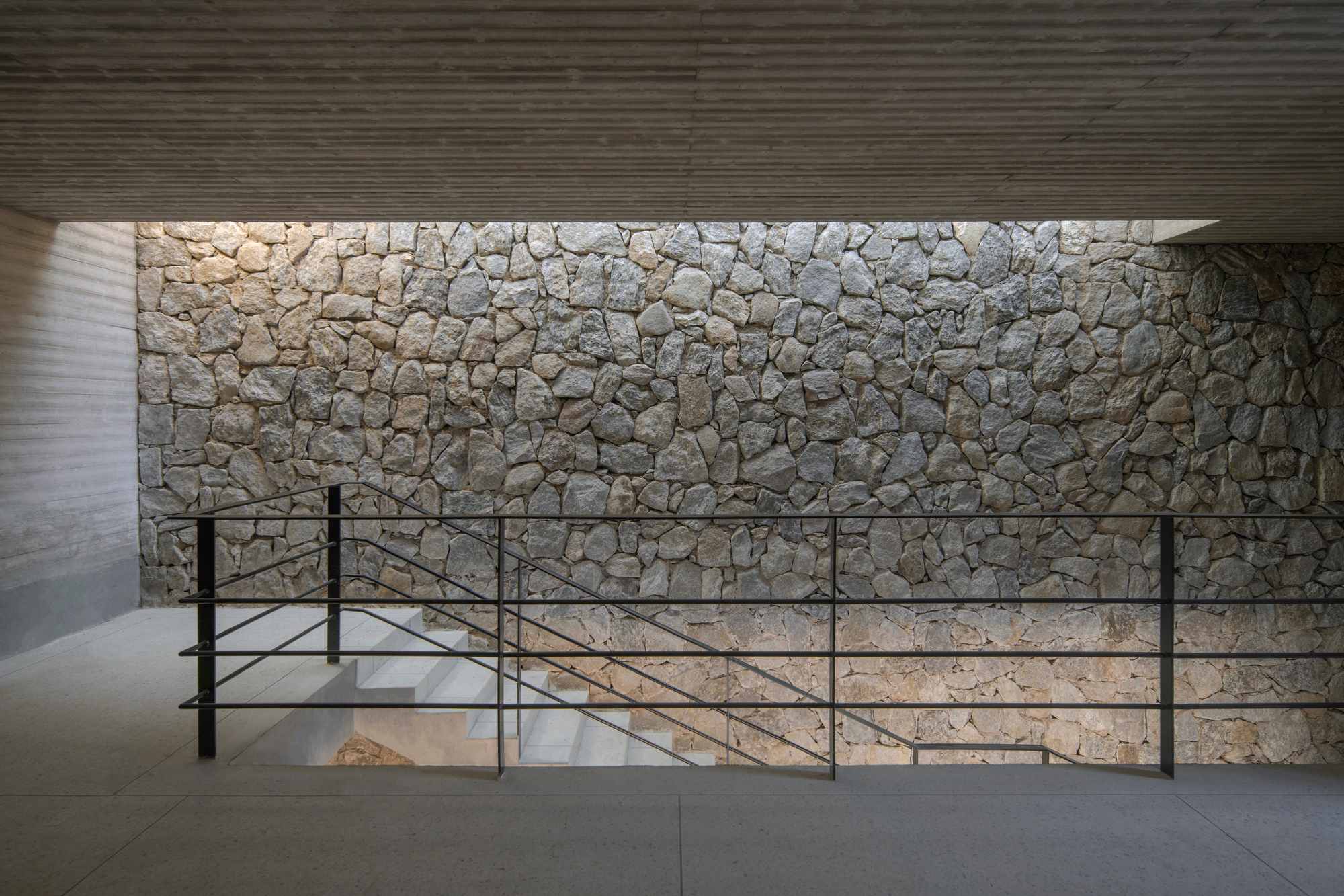 Stone wall lit from above behind a black steel railing and a concrete stair descending below a board-formed ceiling