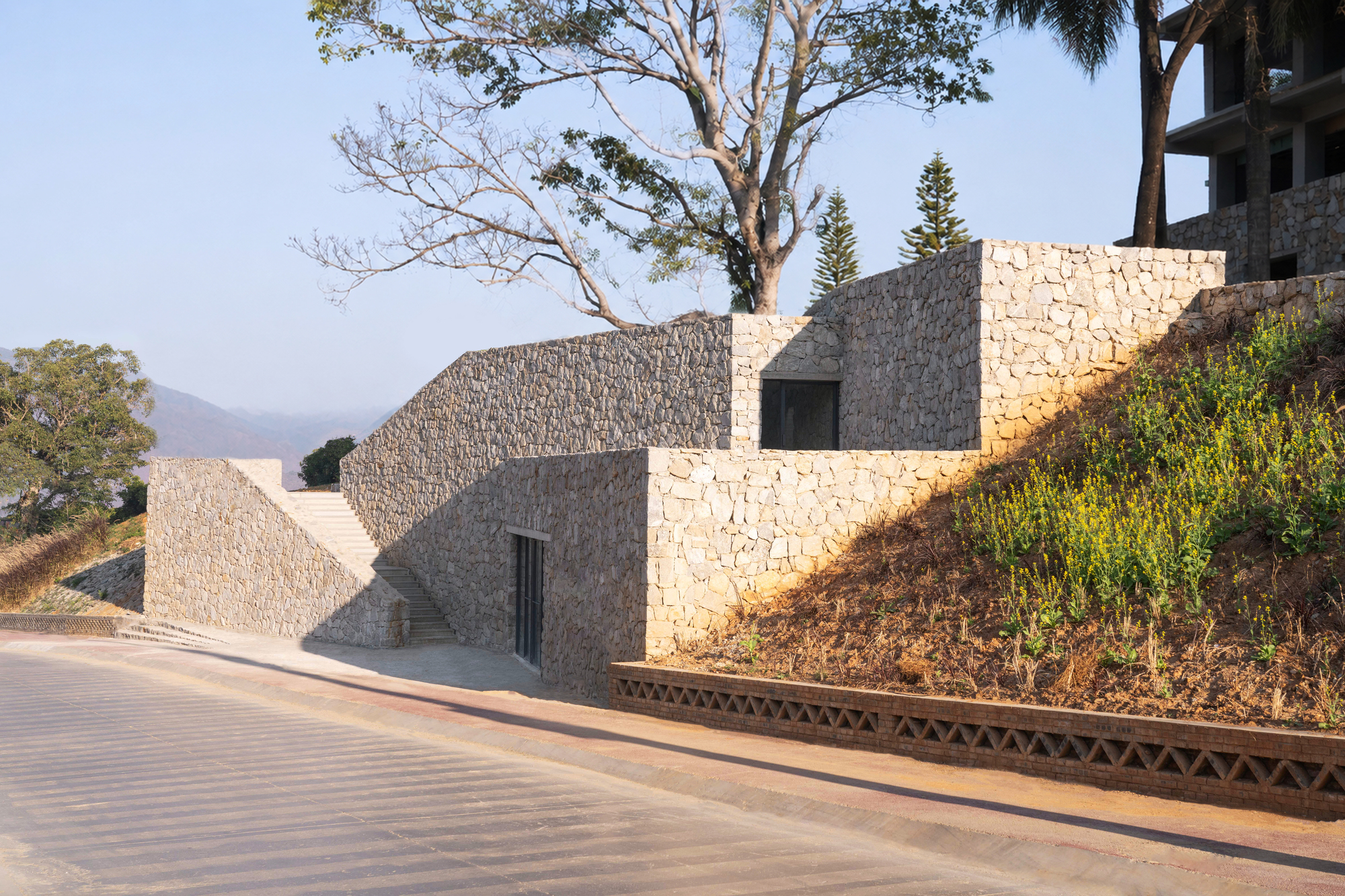 This photograph displays an earth-integrated building embedded into a sloping hillside, constructed from textured light-colored stone masonry. Terraced monolith