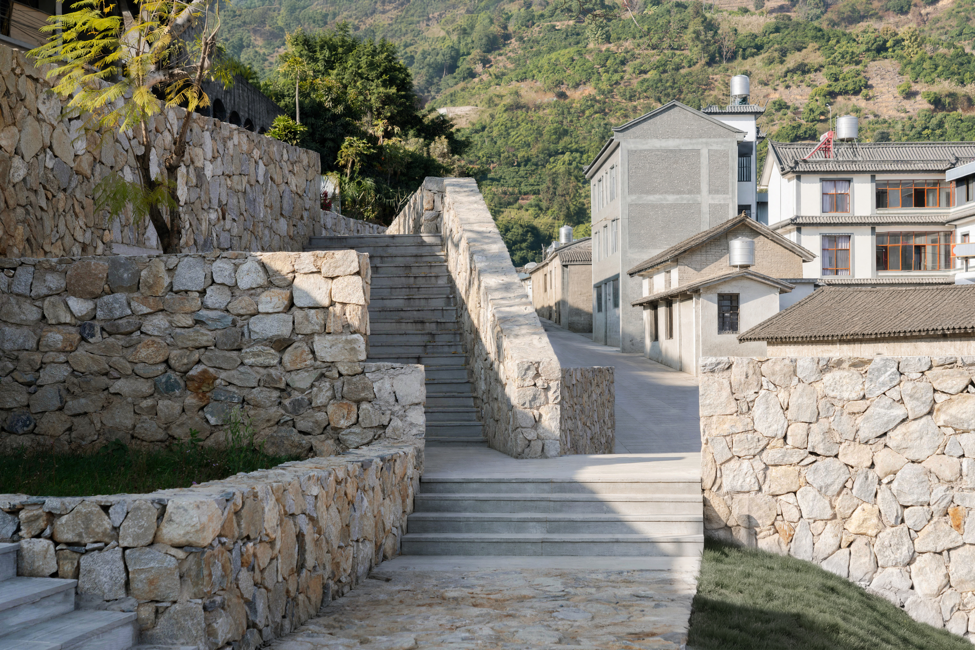 This photograph captures a stepped outdoor circulation area in a hillside settlement, framed by prominent textured stone retaining walls. Clean-lined concrete s