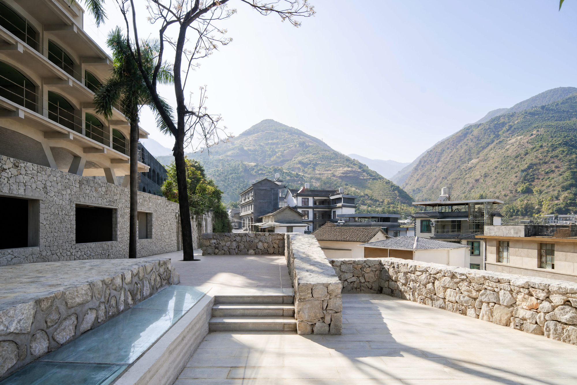 An expansive outdoor stone terrace and observation deck overlooks a valley settlement nestled against dramatic mountain peaks. Built using textured ashlar and r