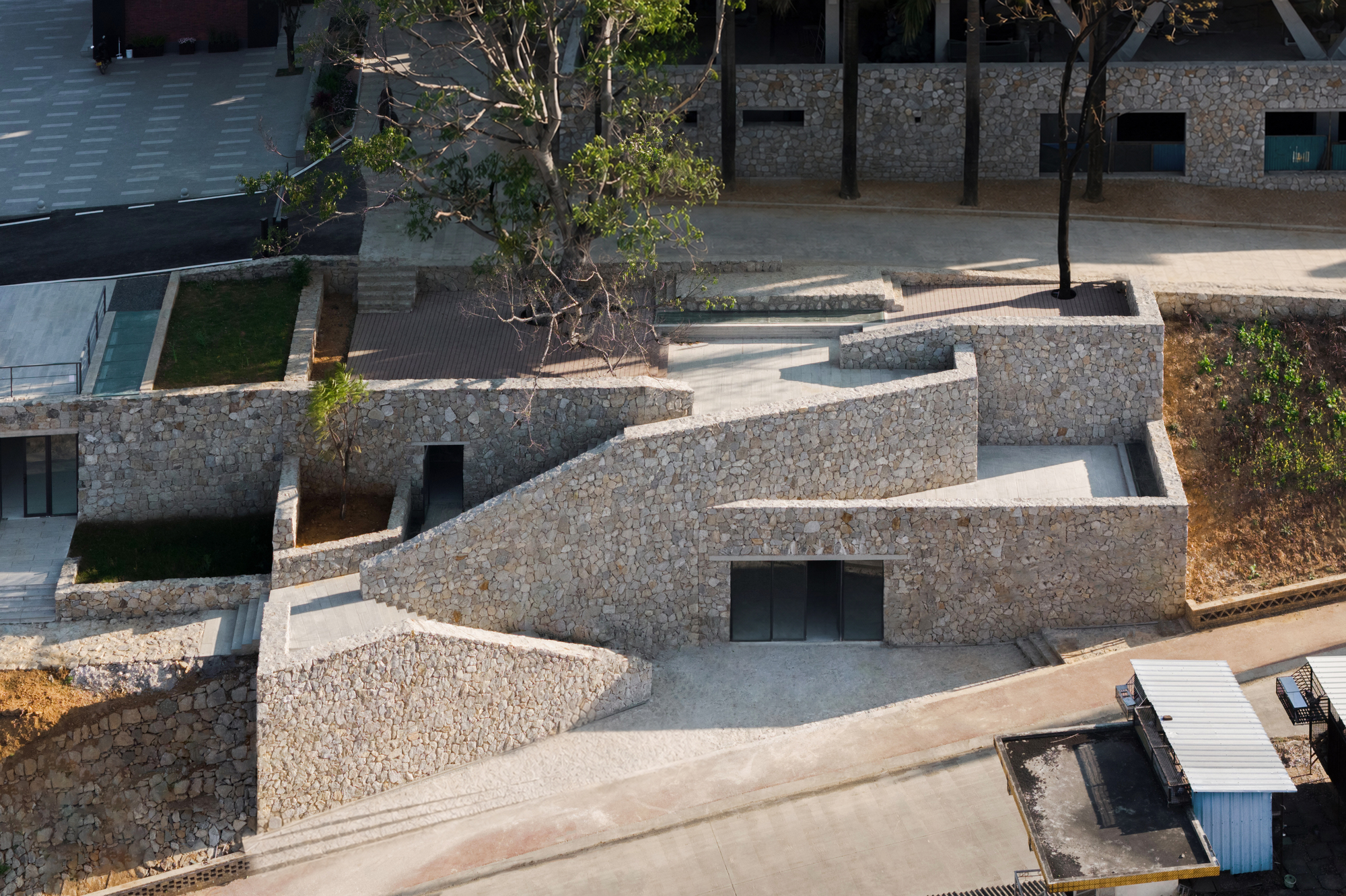 Aerial view of terraced rubble stone walls with a ramp, dark glazed doors and a leafy tree in afternoon sun