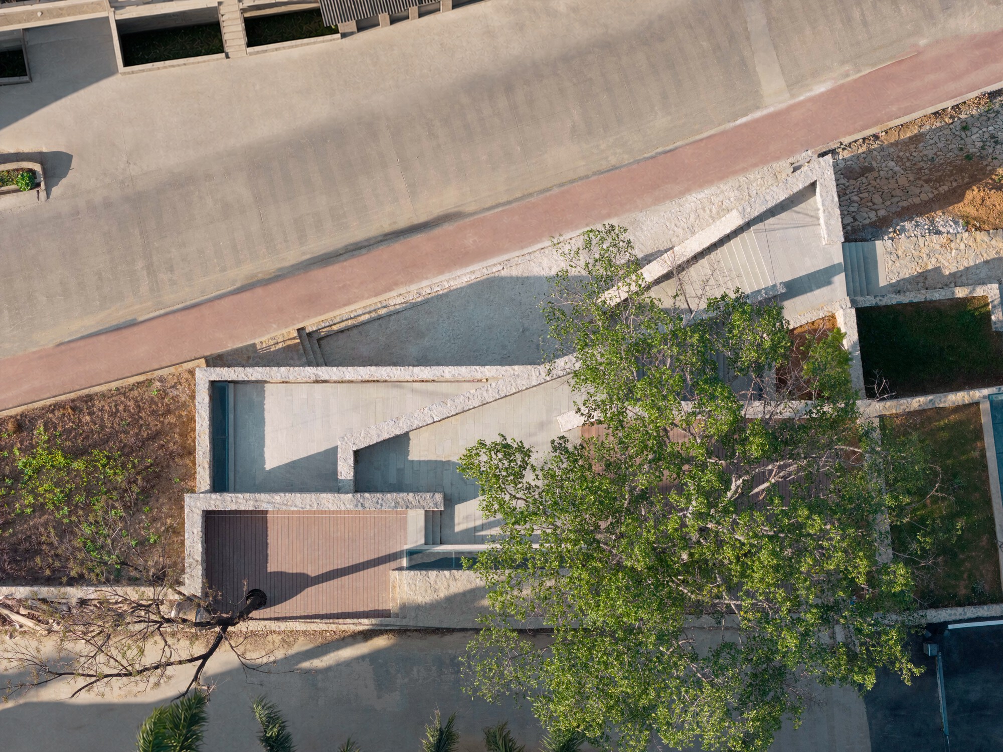 Top-down view of stepped stone-edged terraces with a timber deck and a large tree canopy beside a brick-red path