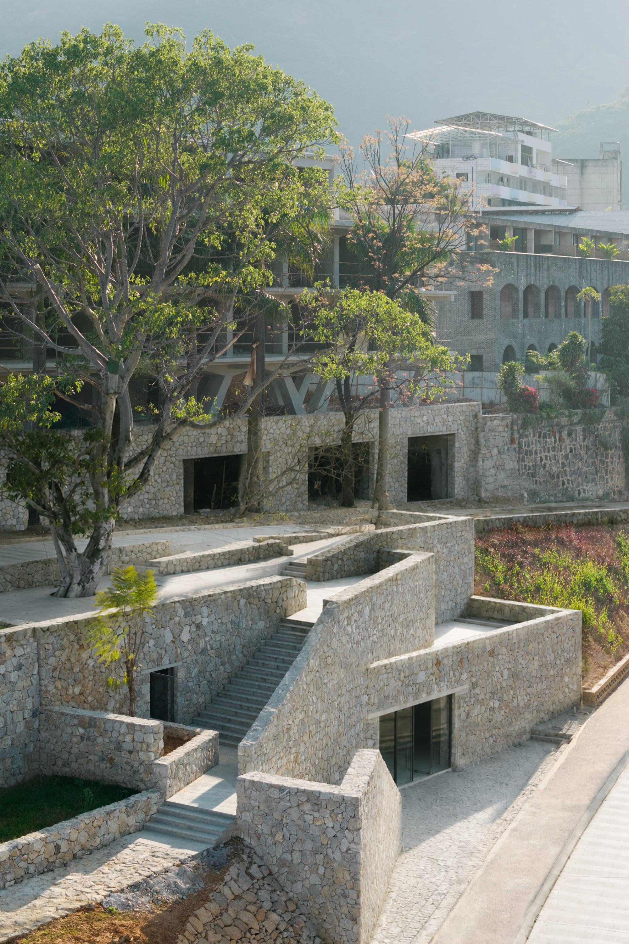 Stone retaining walls and a narrow staircase stepping up toward a hillside building, with mature trees in soft haze