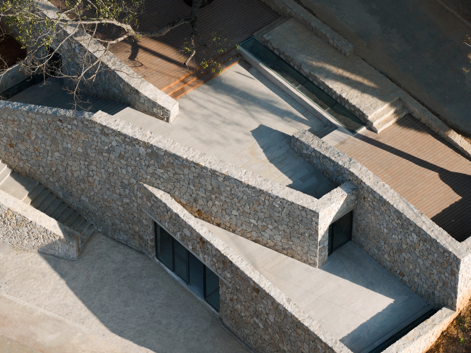 Close aerial view of angled rubble stone walls, glazed openings and a timber deck casting long shadows in low sun