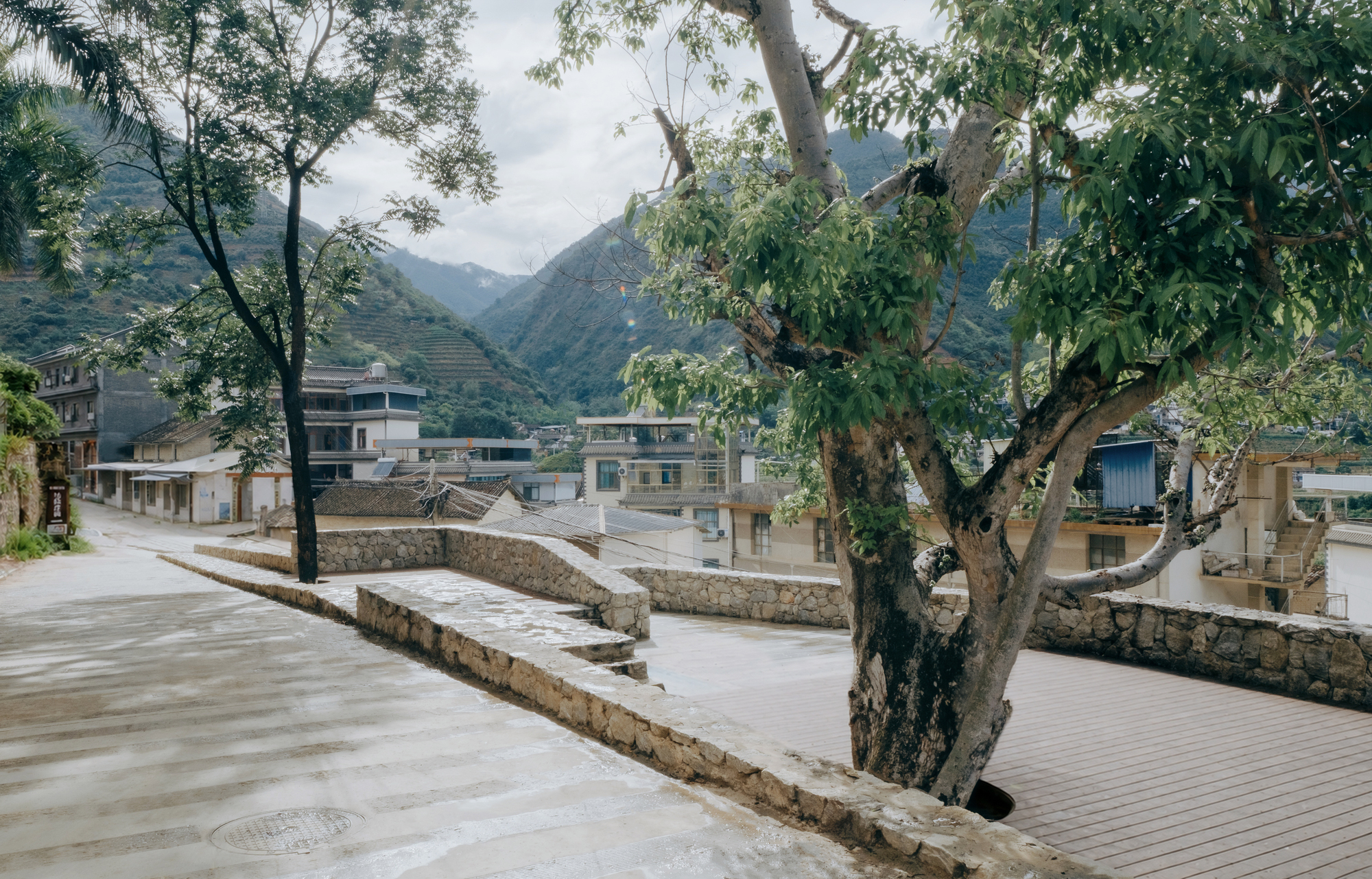 Paved terrace with low stone seating walls and a mature tree, looking toward village houses and terraced mountains under overcast sky