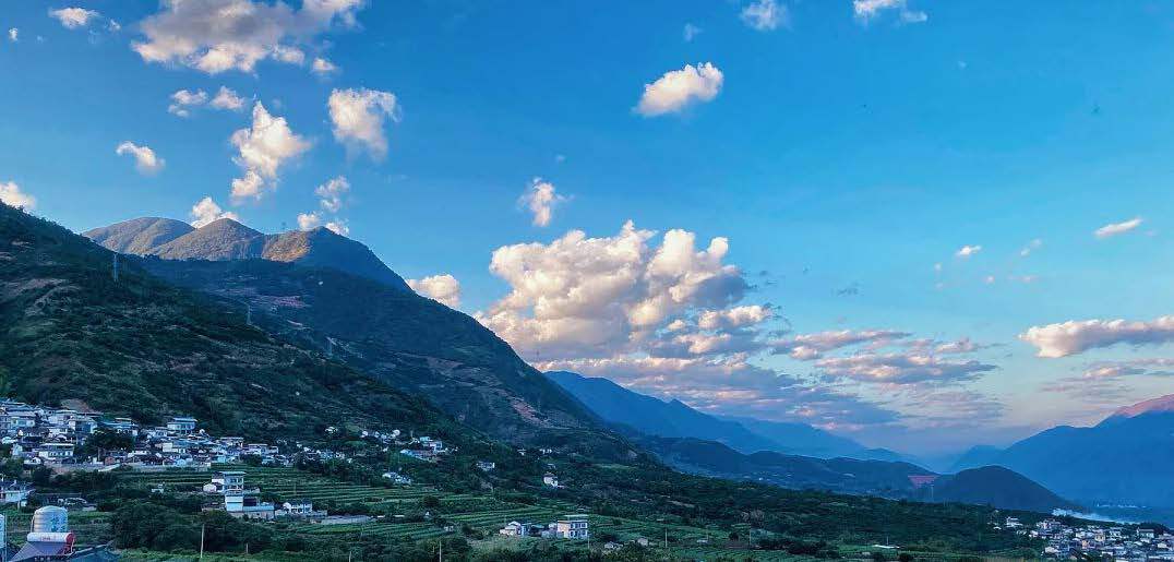 Village houses and terraced orchards on a mountain slope under a blue sky with scattered cumulus clouds