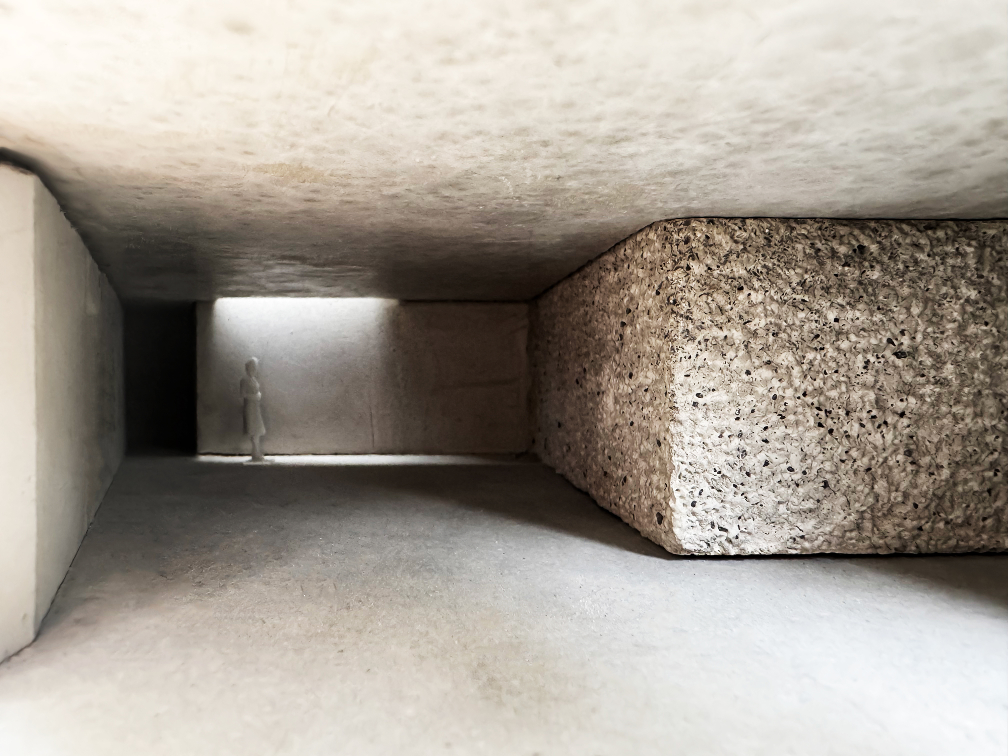 Low view down a model corridor with a pitted concrete block on the right and a lone figure lit at the far end
