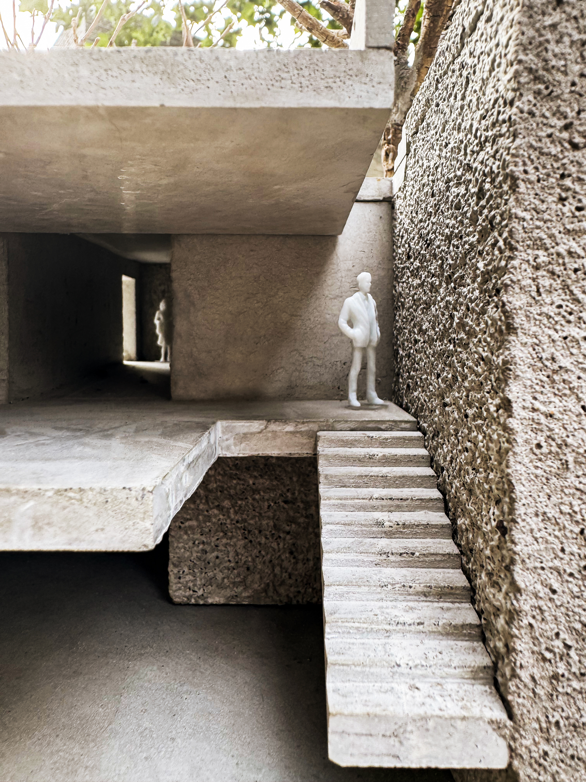 Model detail of a floating concrete stair beside a porous stone wall, with a standing figure and leafy branches overhead