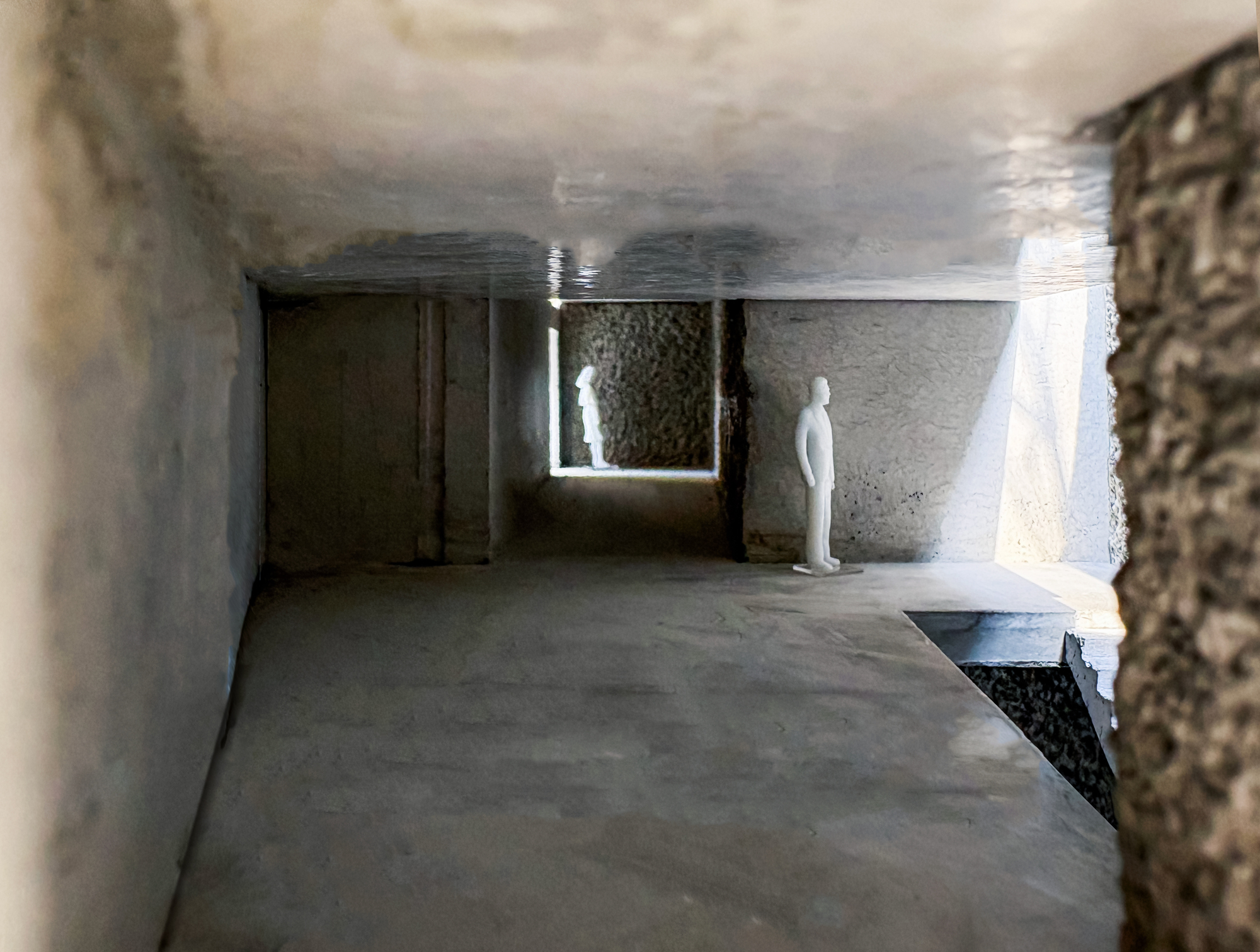 Model interior with two white figures in a dim concrete room, daylight entering through a framed opening and a side light well