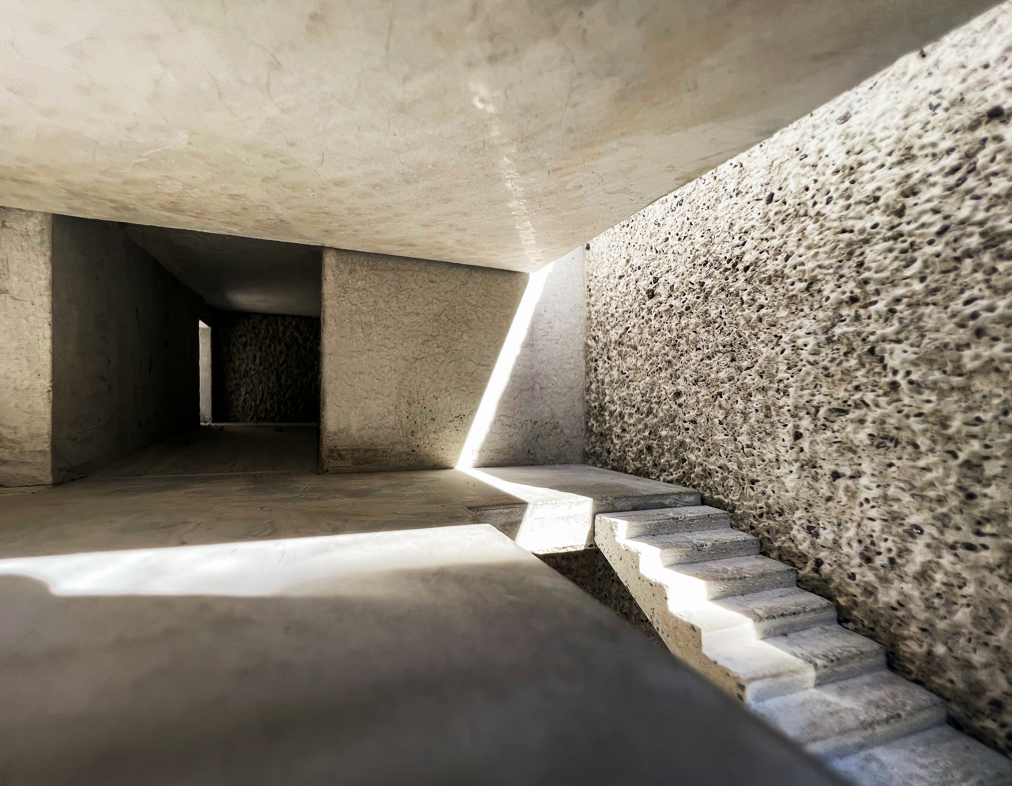 Model interior of a rough concrete stair beside a pitted wall, with a sharp shaft of sunlight cutting across the floor