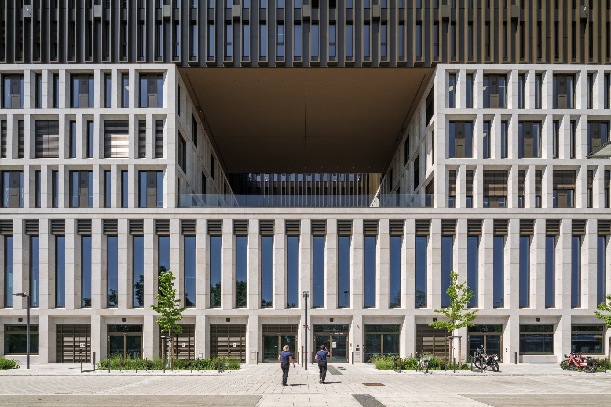 Central recess in the stone grid facade with tall ground-floor windows and two pedestrians crossing the paved square in sunlight