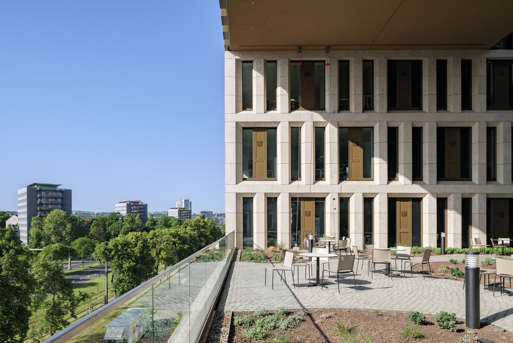 Roof terrace with café tables and planted beds beside the stone facade, overlooking treetops and a glass balustrade
