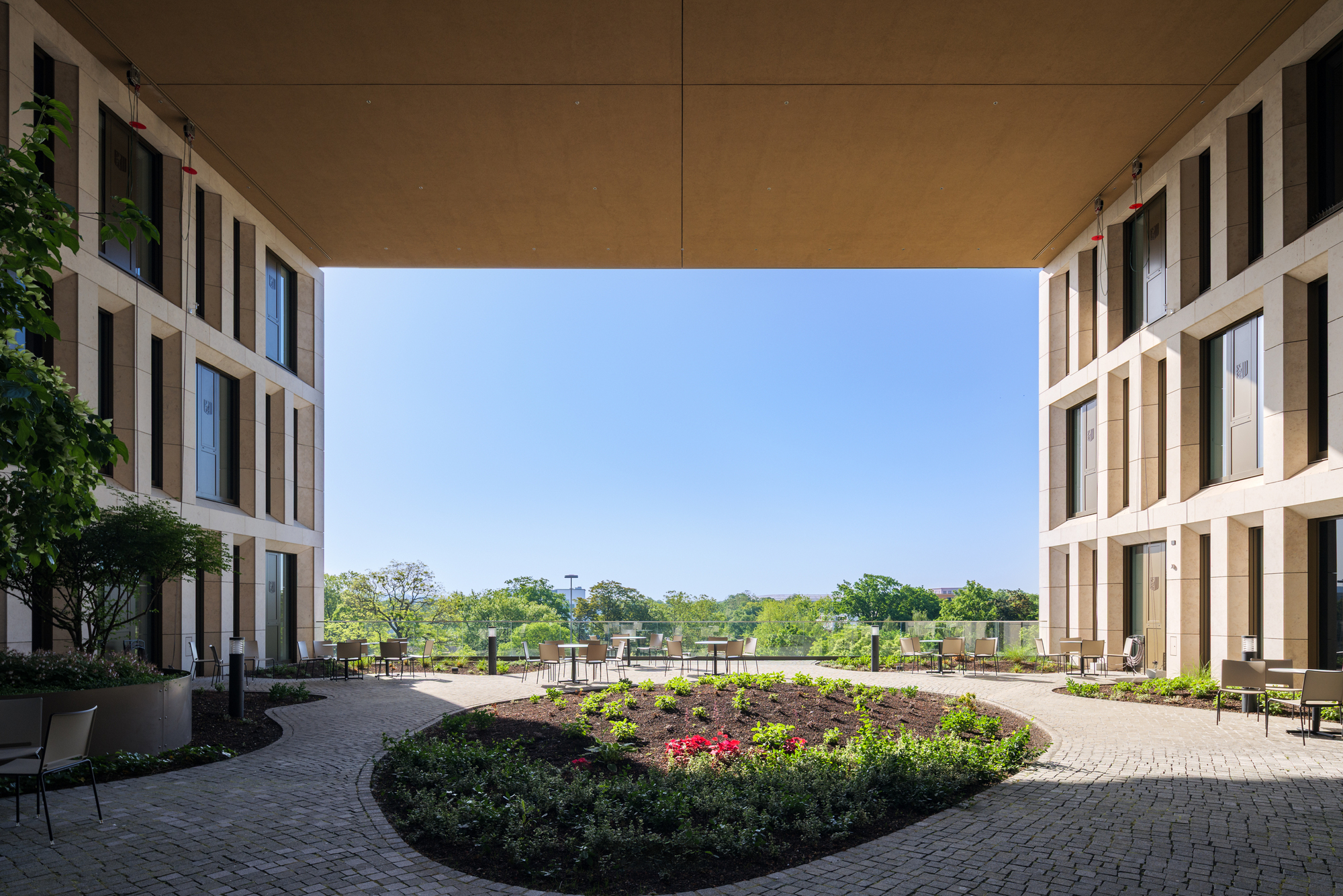 Covered passage opening onto a terrace with a curved planted bed, brick paving and tables under a timber-toned soffit