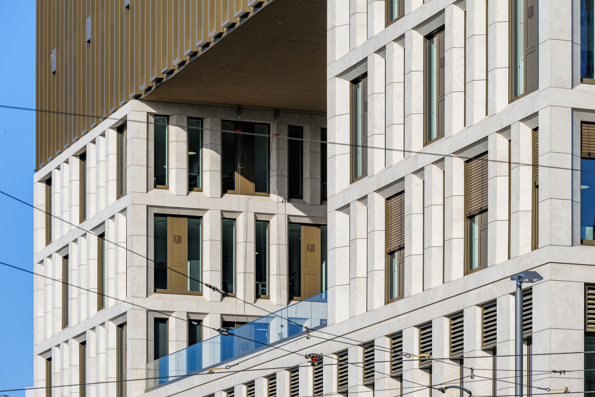 Angled view of the stone facade with timber shutters and a glass balustrade, crossed by tram wires against blue sky