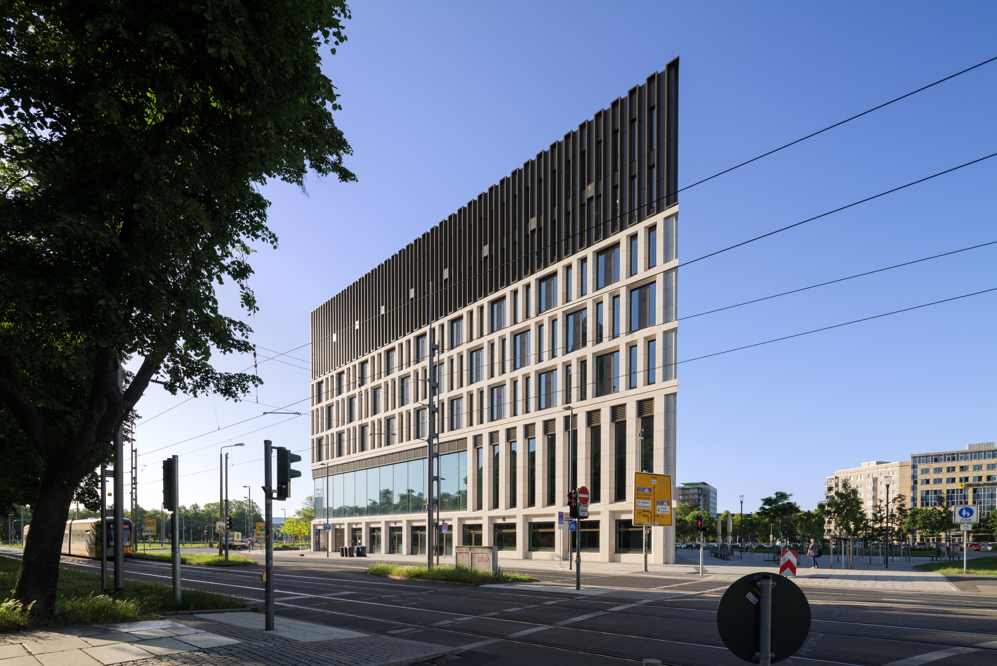Corner of the stone-framed facade with a dark vertical-fin upper storey, seen across tram tracks under a clear blue sky