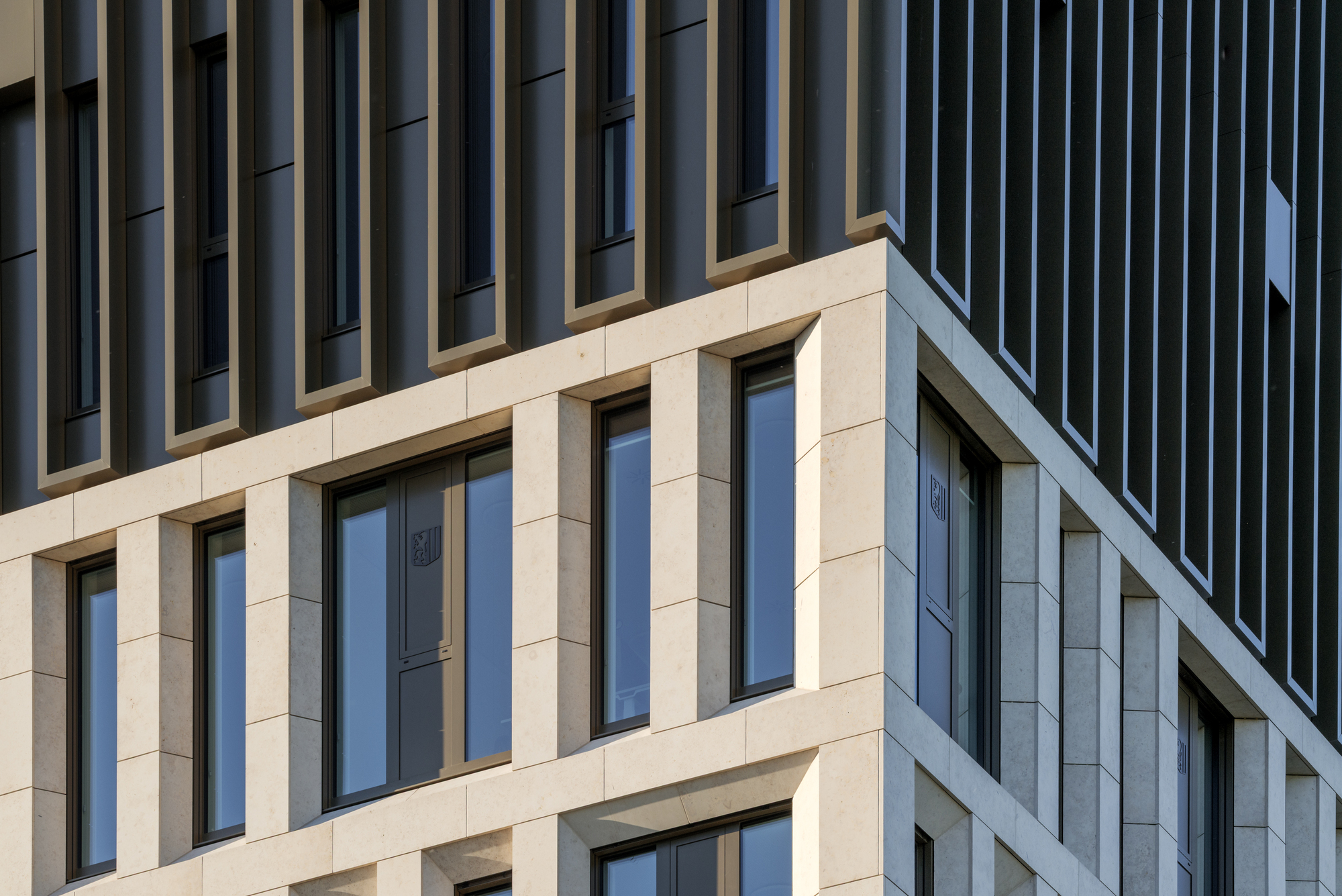 Close-up of the pale stone grid and recessed windows meeting the dark finned upper facade in low sun