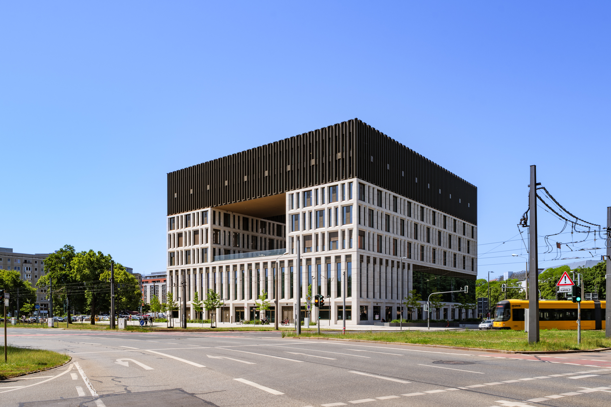 Cube-shaped block with a cut-out corner and dark fin cladding, a yellow tram passing on the street at right