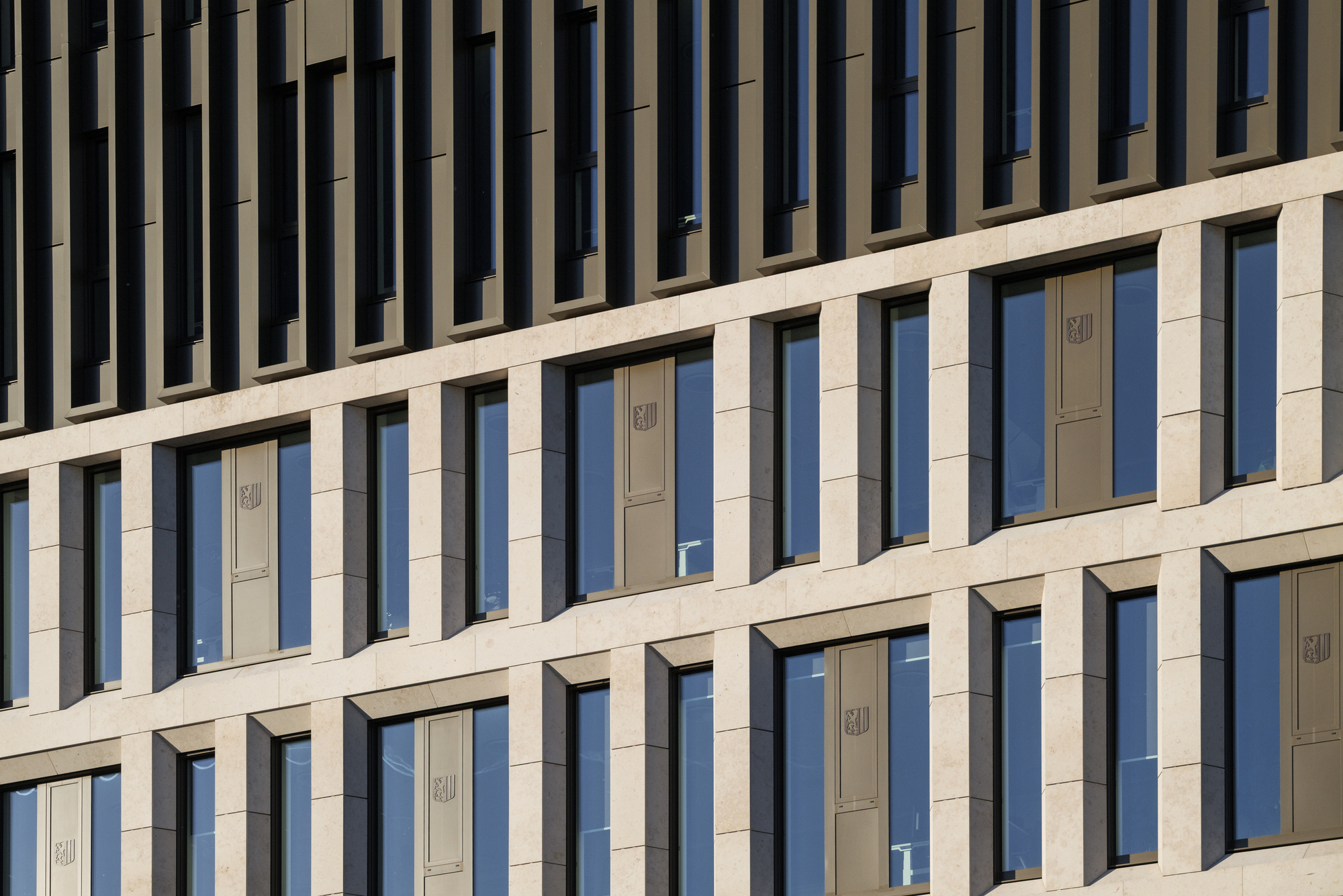 Close-up of limestone facade with deep window reveals and bronze-toned shutters bearing a small crest, in low raking sunlight