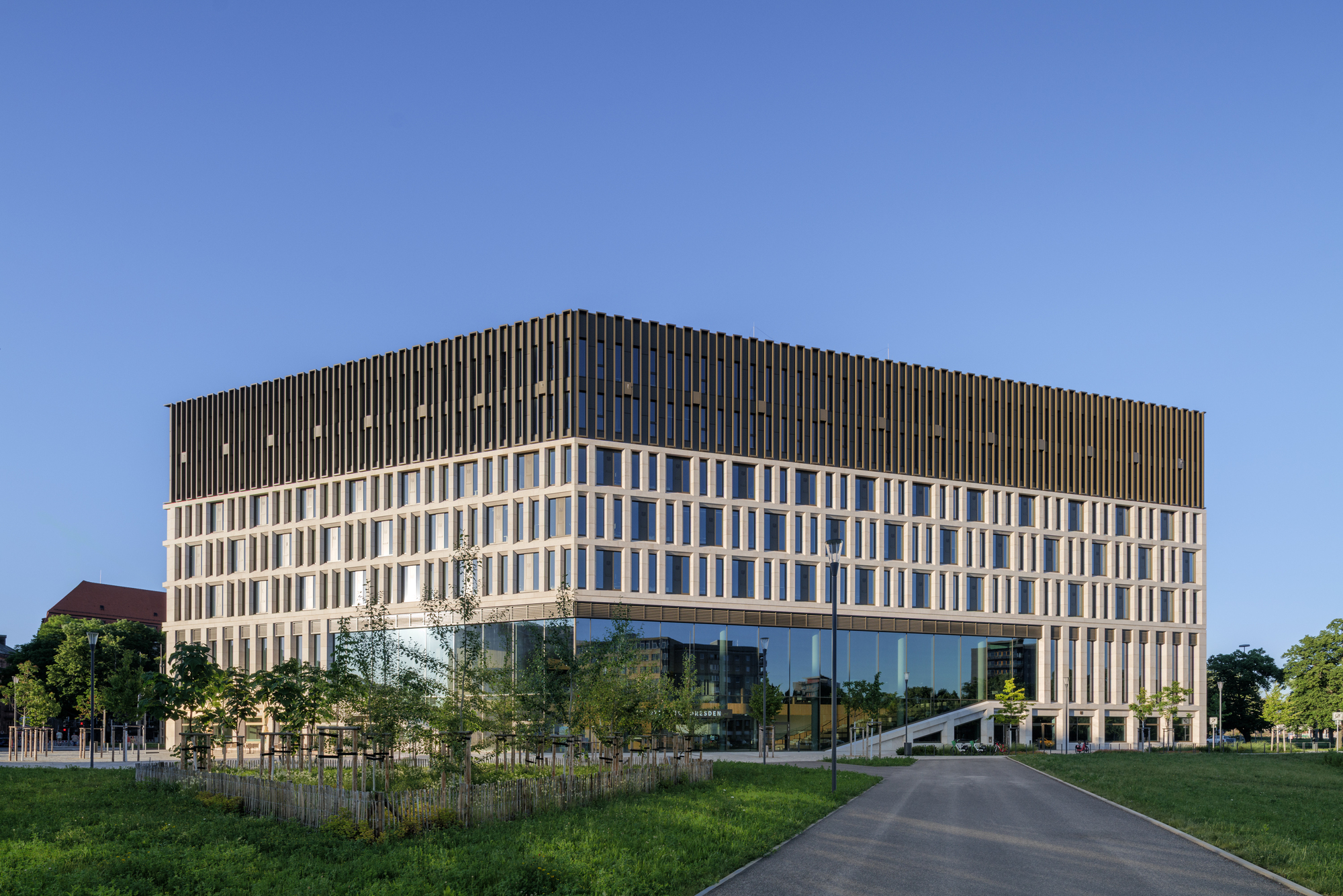 Corner view of the cube-shaped building with a vertical-fin bronze upper level above a glazed base, seen across a lawn under clear blue sky