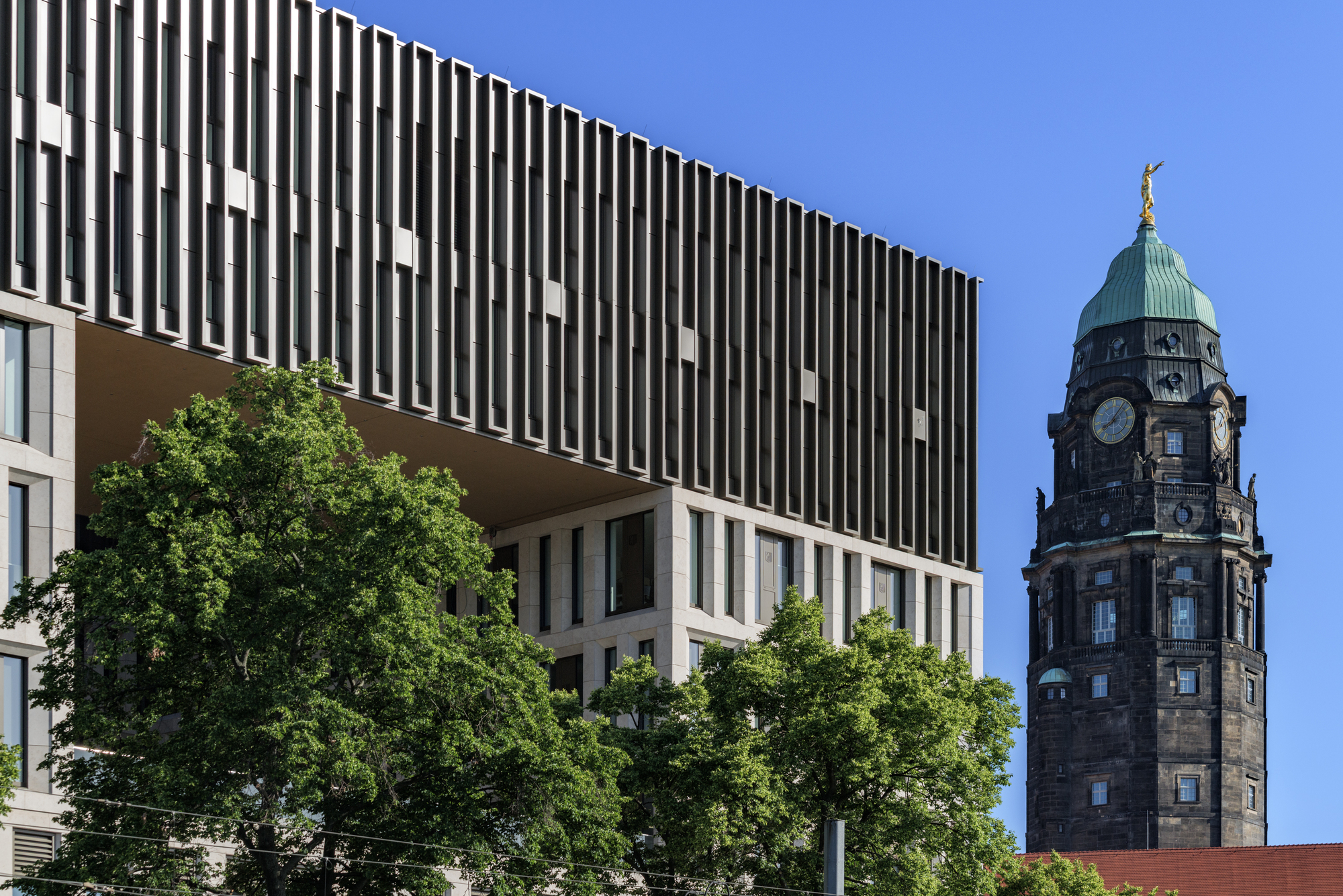 Overhanging fin-clad upper volume beside a dark clock tower with a green copper dome, framed by summer trees